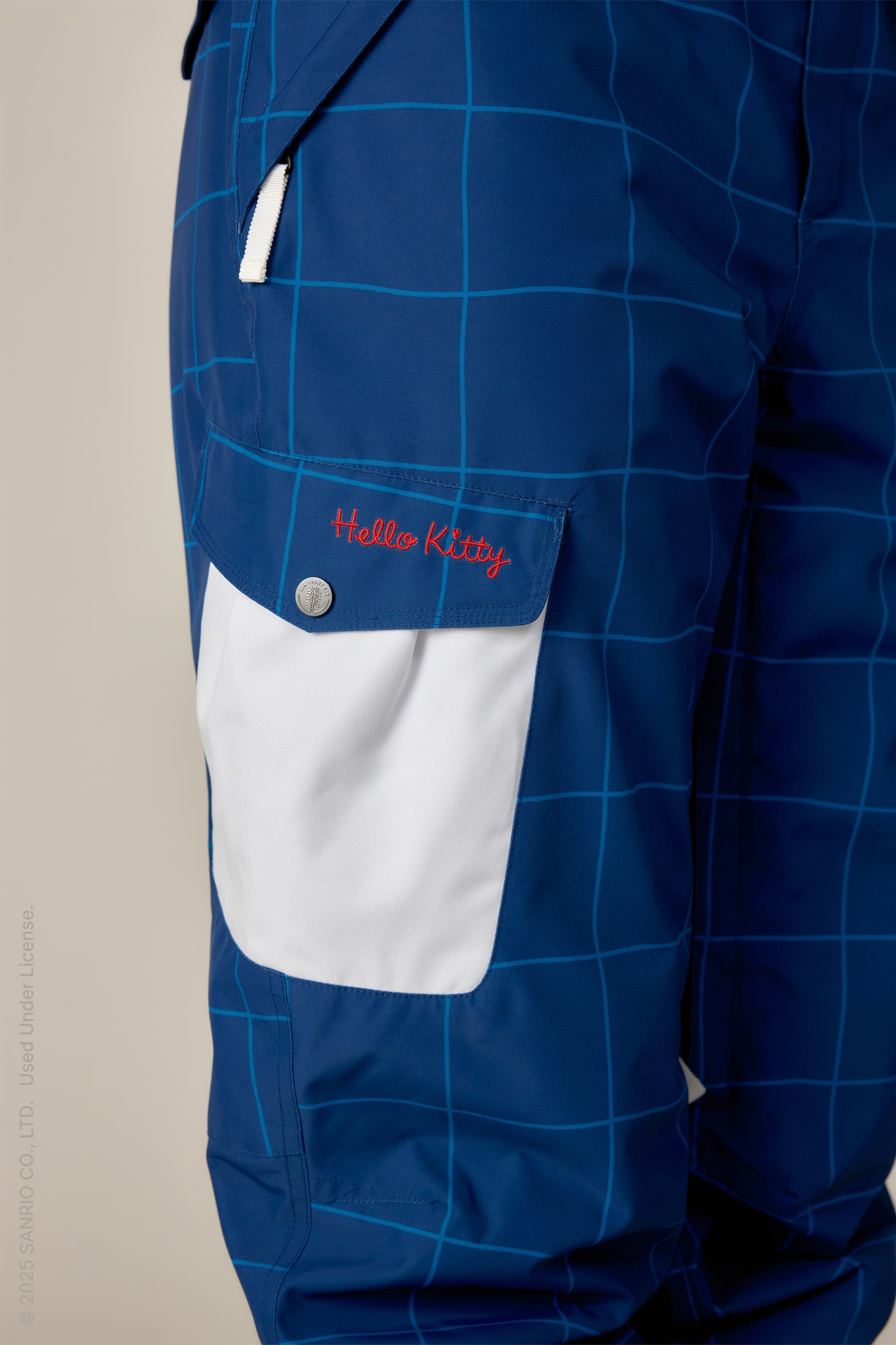 Close-up of the 686 Womens Aura Insulated Cargo Pant in blue with a grid pattern, featuring a white pocket flap embroidered with red Hello Kitty text and a small white zipper above the pocket.
