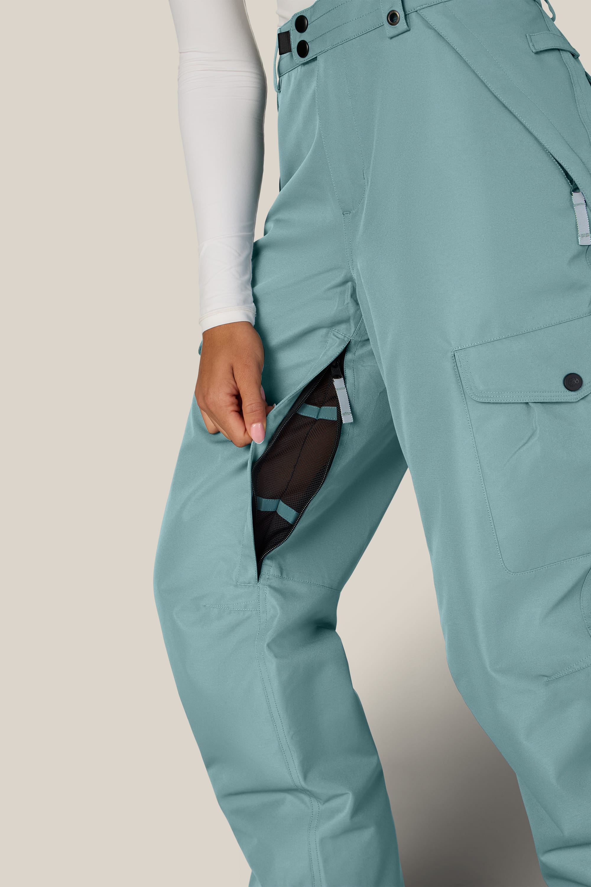 A person wearing the 686 Women’s Aura™ Insulated Cargo Pant in teal and a white long-sleeve top unzips a side vent on the pants, revealing black mesh fabric inside.
