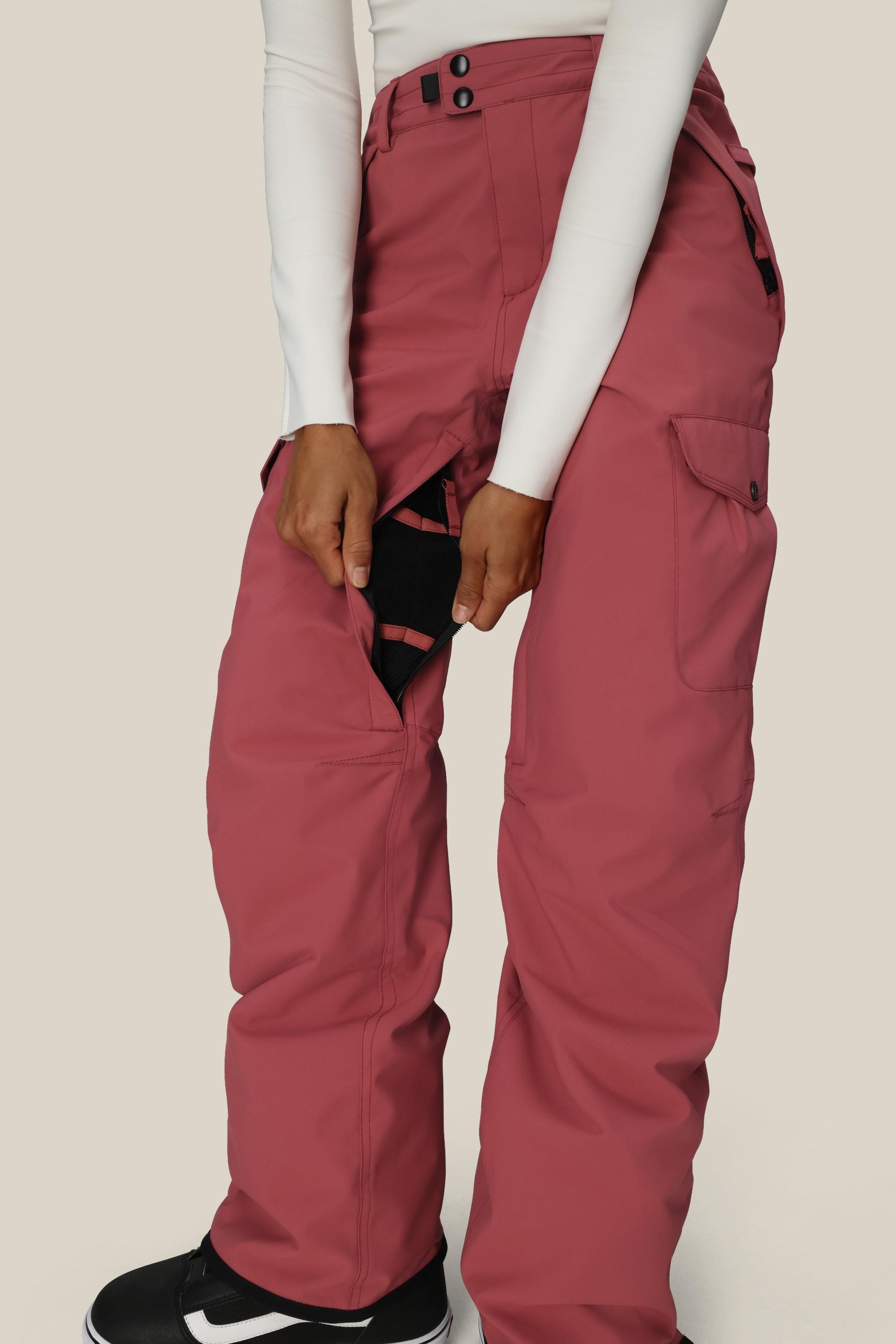 Wearing 686 Womens Aura Insulated Cargo Pant, a person in rose-colored waterproof pants and a white long-sleeve top opens a zippered side vent to show the black mesh lining; only their lower body is visible.