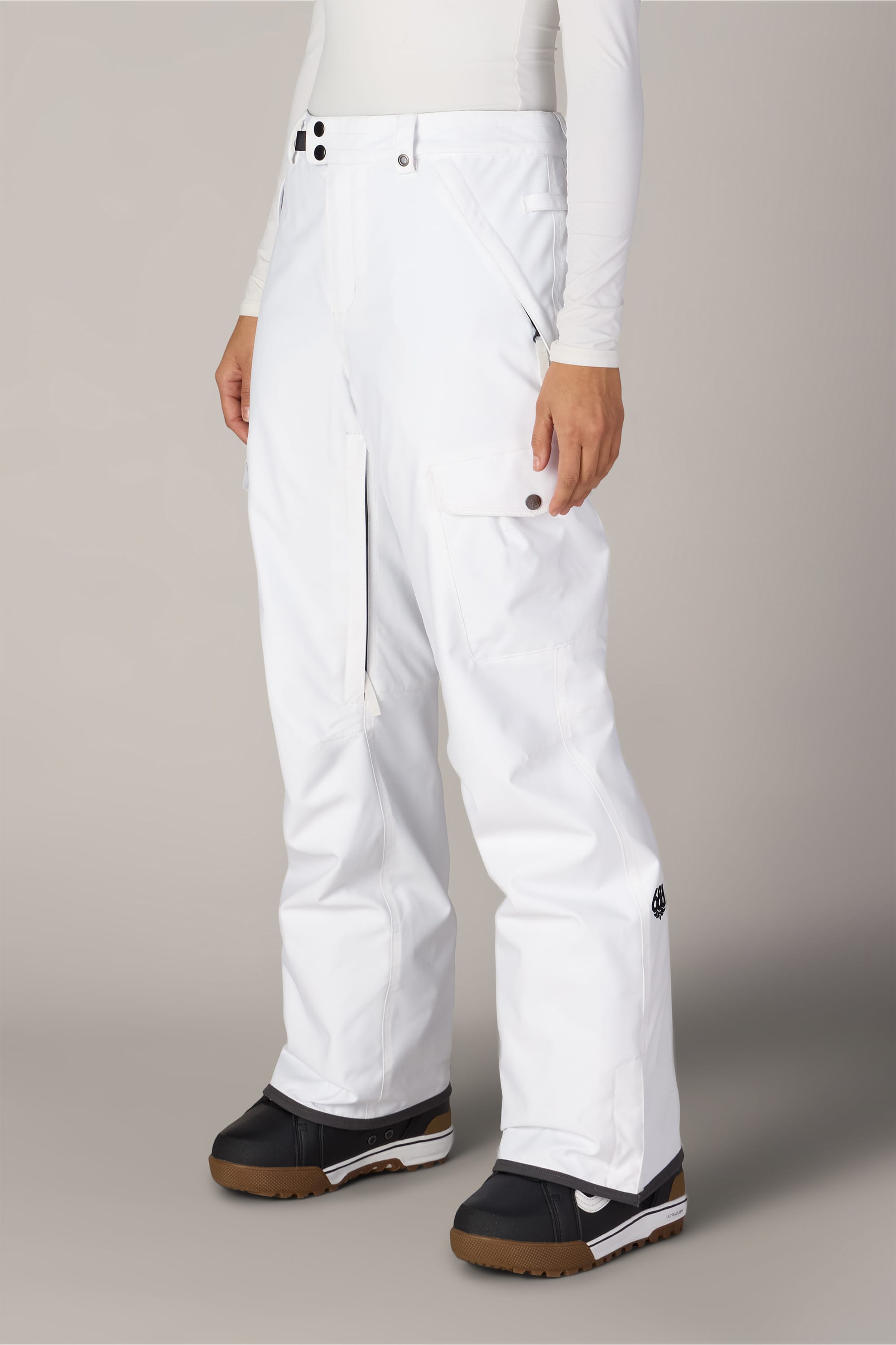 A person wearing the 686 Women’s Aura™ Insulated Cargo Pant by 686, paired with a white long-sleeve shirt and black snow boots, standing against a neutral background.