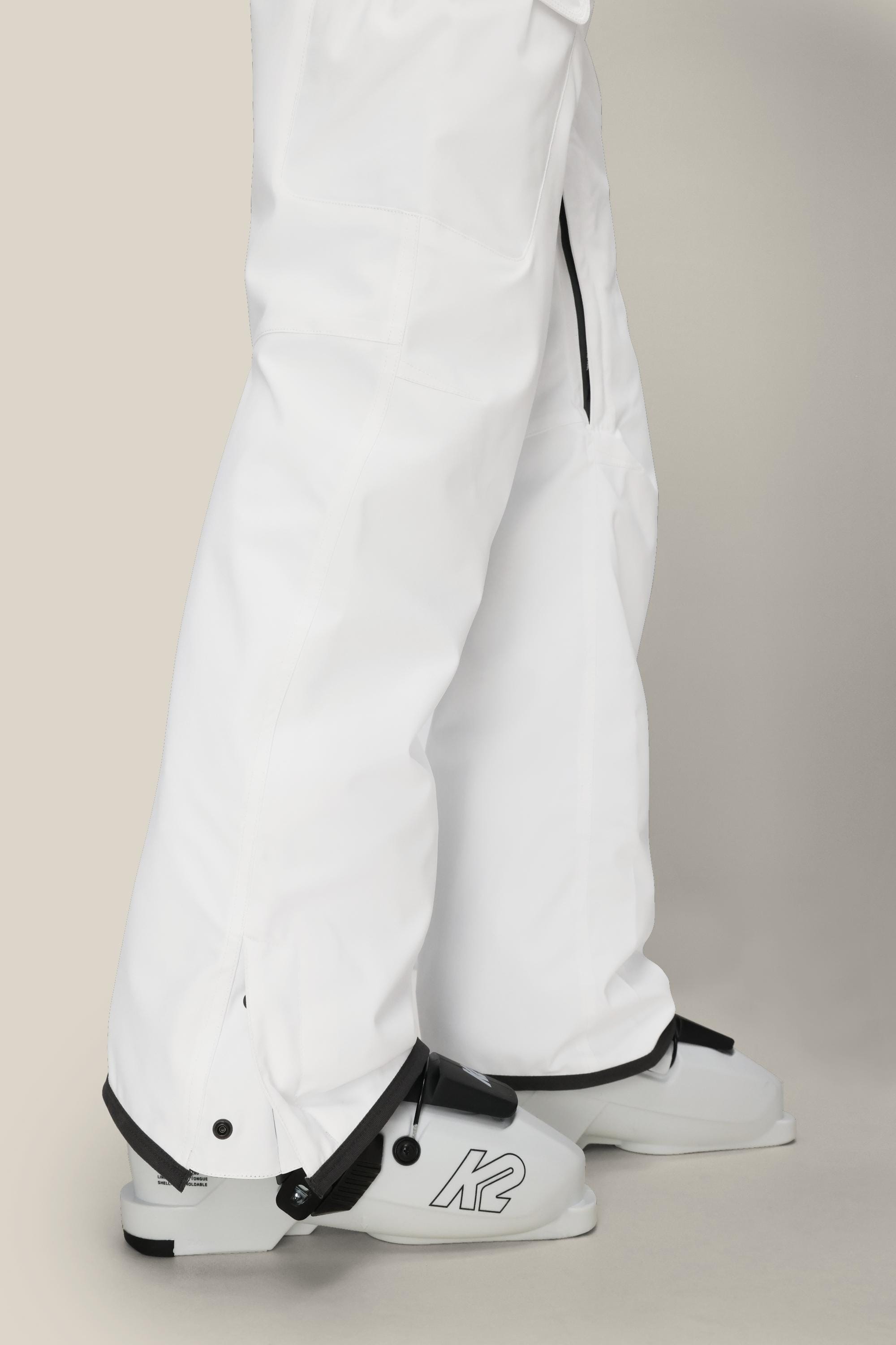A person wearing 686 Womens Aura Insulated Cargo Pant and white ski boots with K2 branding stands against a plain, light-colored background.