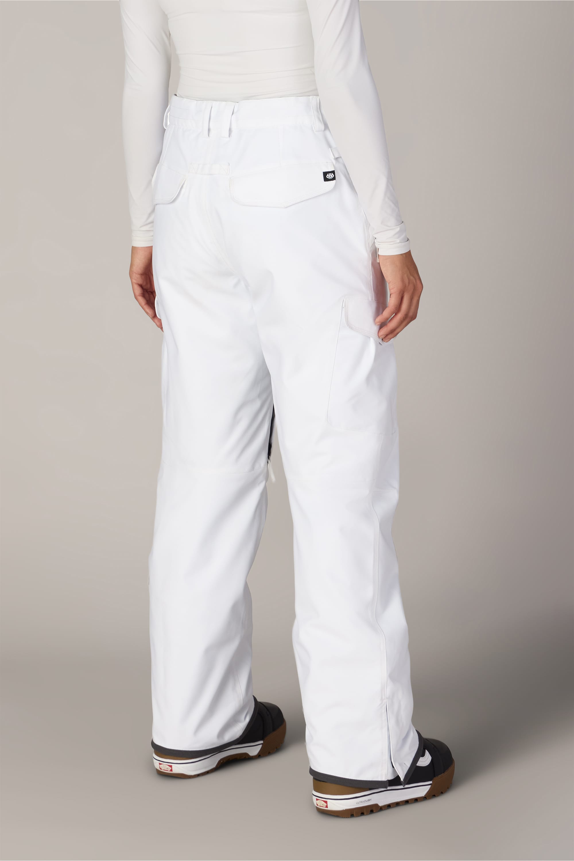 A person is shown from the back, standing against a plain background and wearing the 686 Women’s Aura™ Insulated Cargo Pant by 686 in a loose-fitting white style, paired with a white long-sleeve top and black sneakers with brown soles.