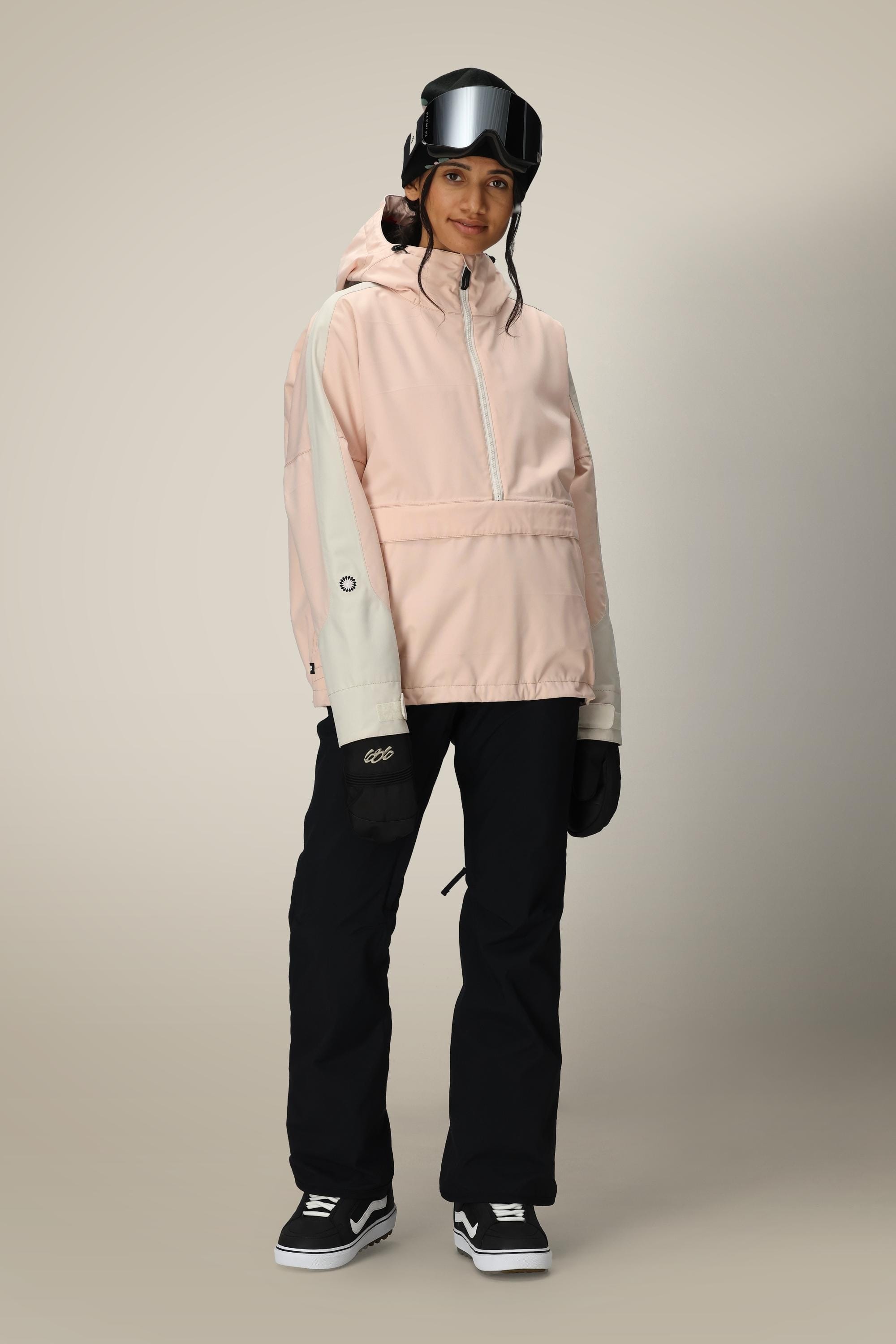 A person is wearing a light pink hooded jacket, the 686 Women’s Standard Shell Pant in black by 686, black gloves, ski goggles, and black sneakers while standing facing forward against a plain background.
