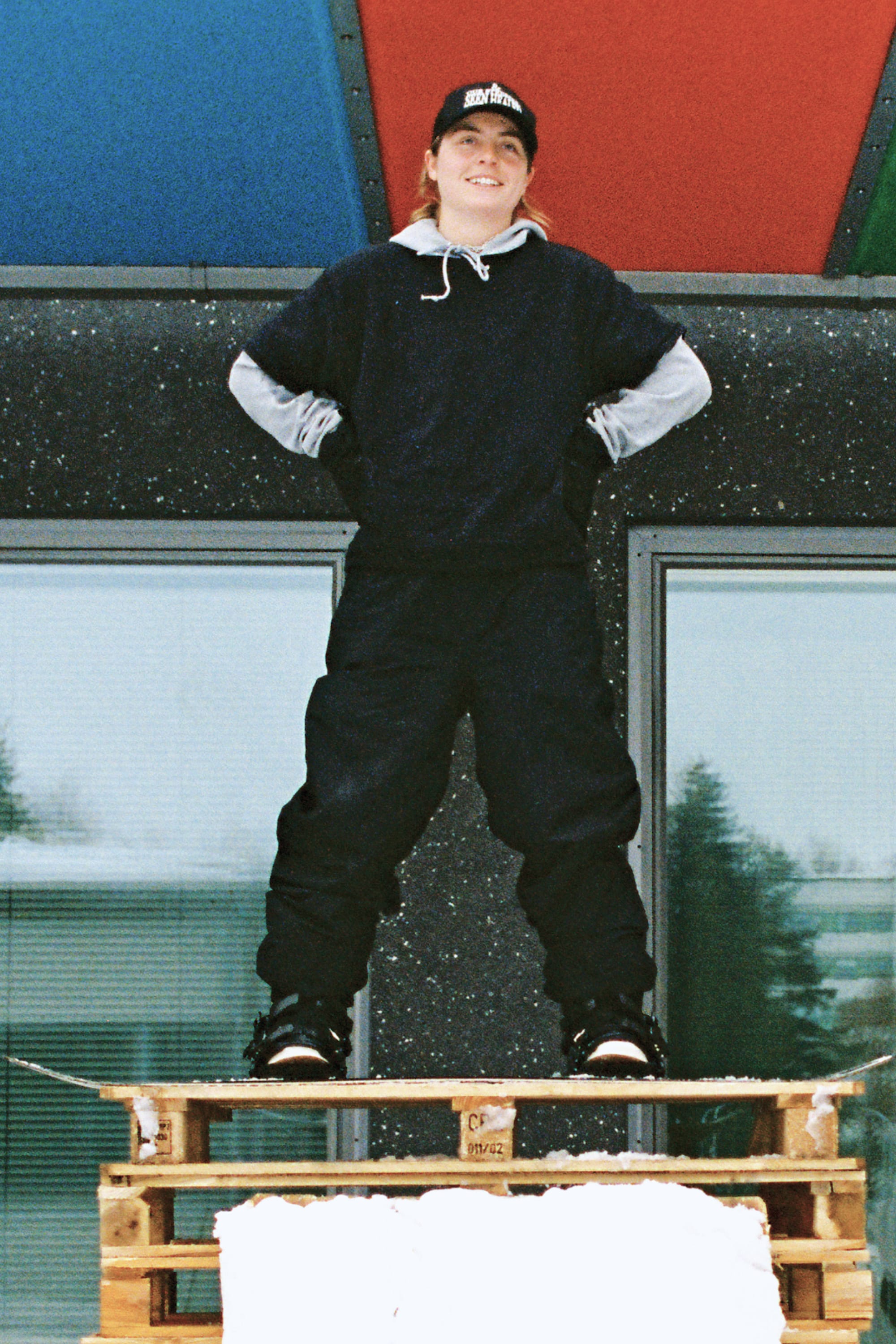 Wearing the 686 Women’s Outline™ Shell Pant by 686, a person dressed in dark winter clothing and a cap stands smiling on stacked snow-covered wooden pallets in front of a building with colorful panels and windows.