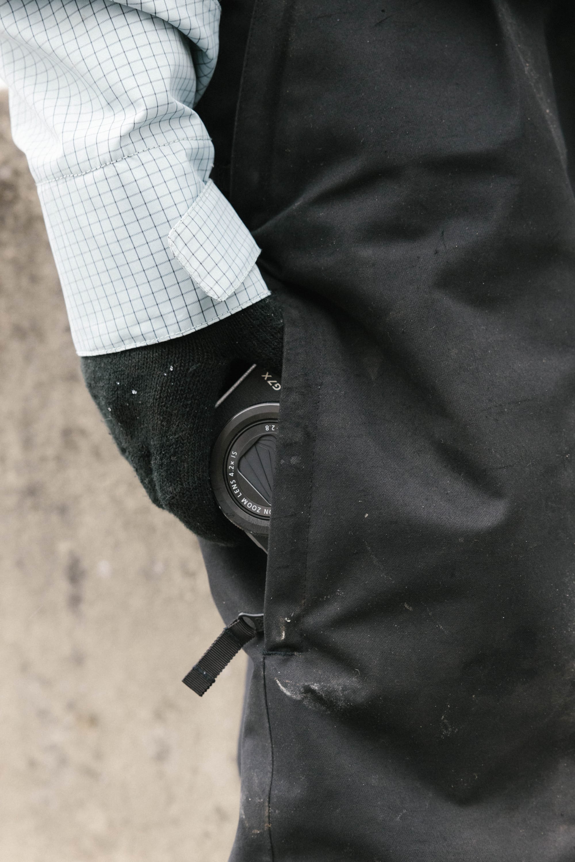 A person wearing a blue checkered shirt and black gloves has a black camera lens partially tucked into the pocket of their 686 Women's Outline™ Shell Pant by 686. The background is blurred and neutral-toned.
