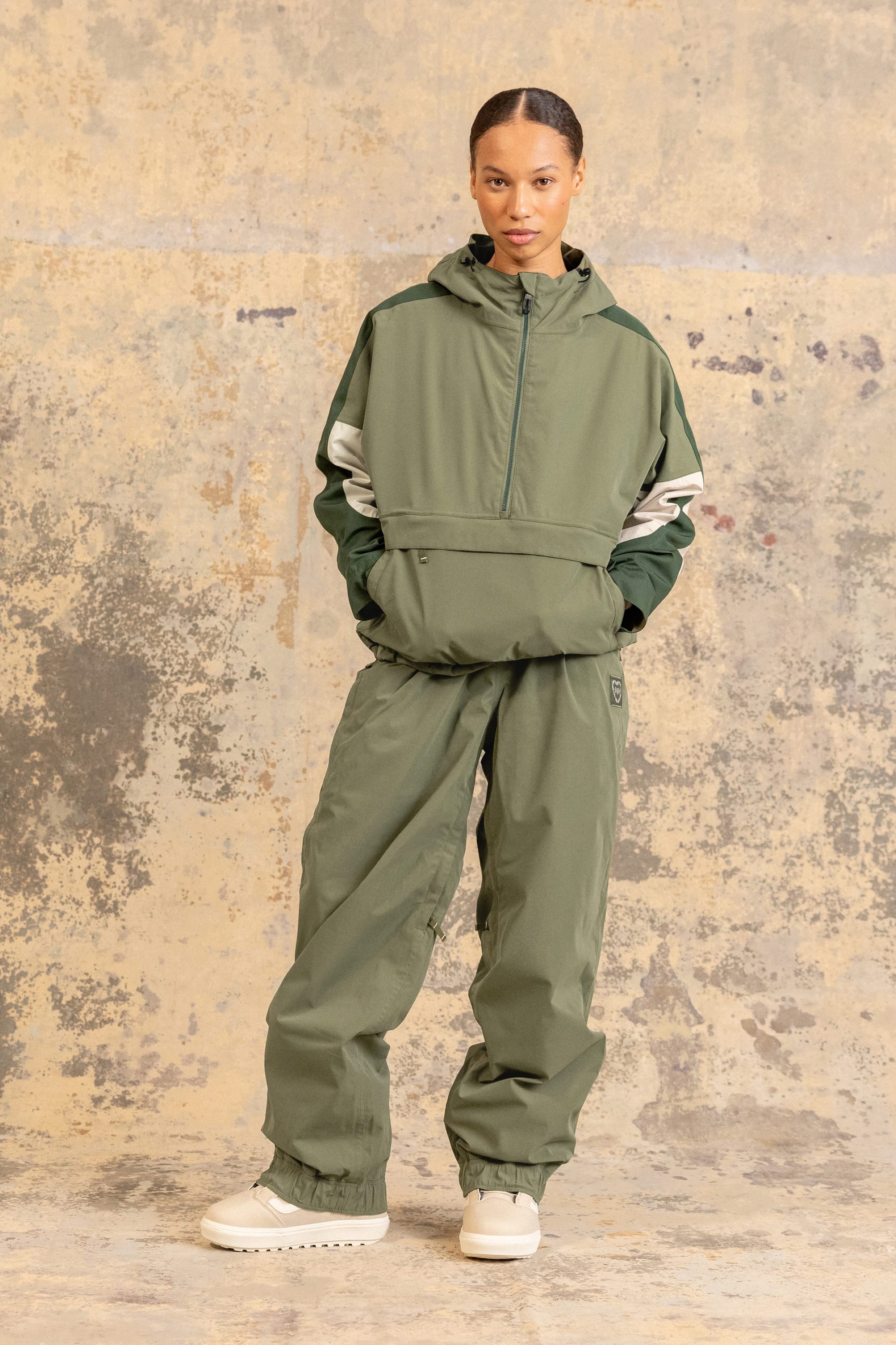 A woman stands against a textured wall wearing the 686 Women’s Outline™ Shell Pant in olive green, paired with a matching hooded jacket and white chunky shoes. She has her hands in her pockets and looks directly at the camera.