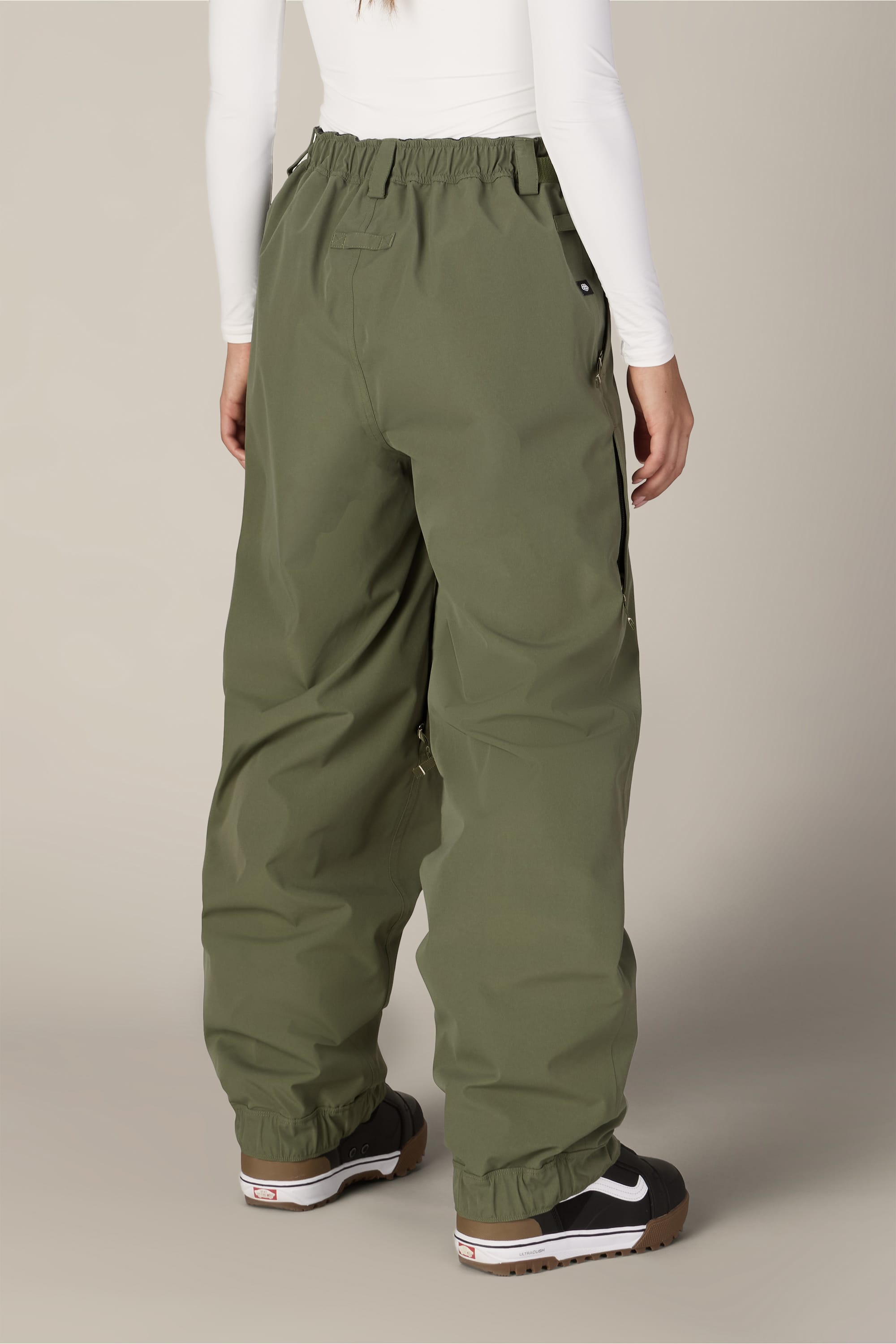 A person is shown from the back standing against a plain background, wearing the 686 Women’s Outline™ Shell Pant in olive green by 686, paired with black and white sneakers and a white long-sleeve top.
