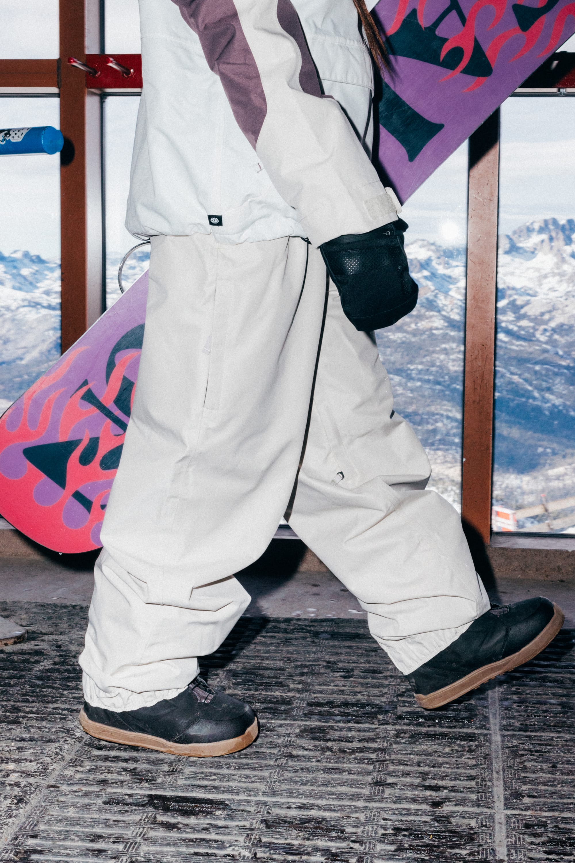 Wearing the 686 Women’s Outline™ Shell Pant by 686, a person stands indoors with black gloves and boots, holding a snowboard as snowy mountains are visible through the window in the background.