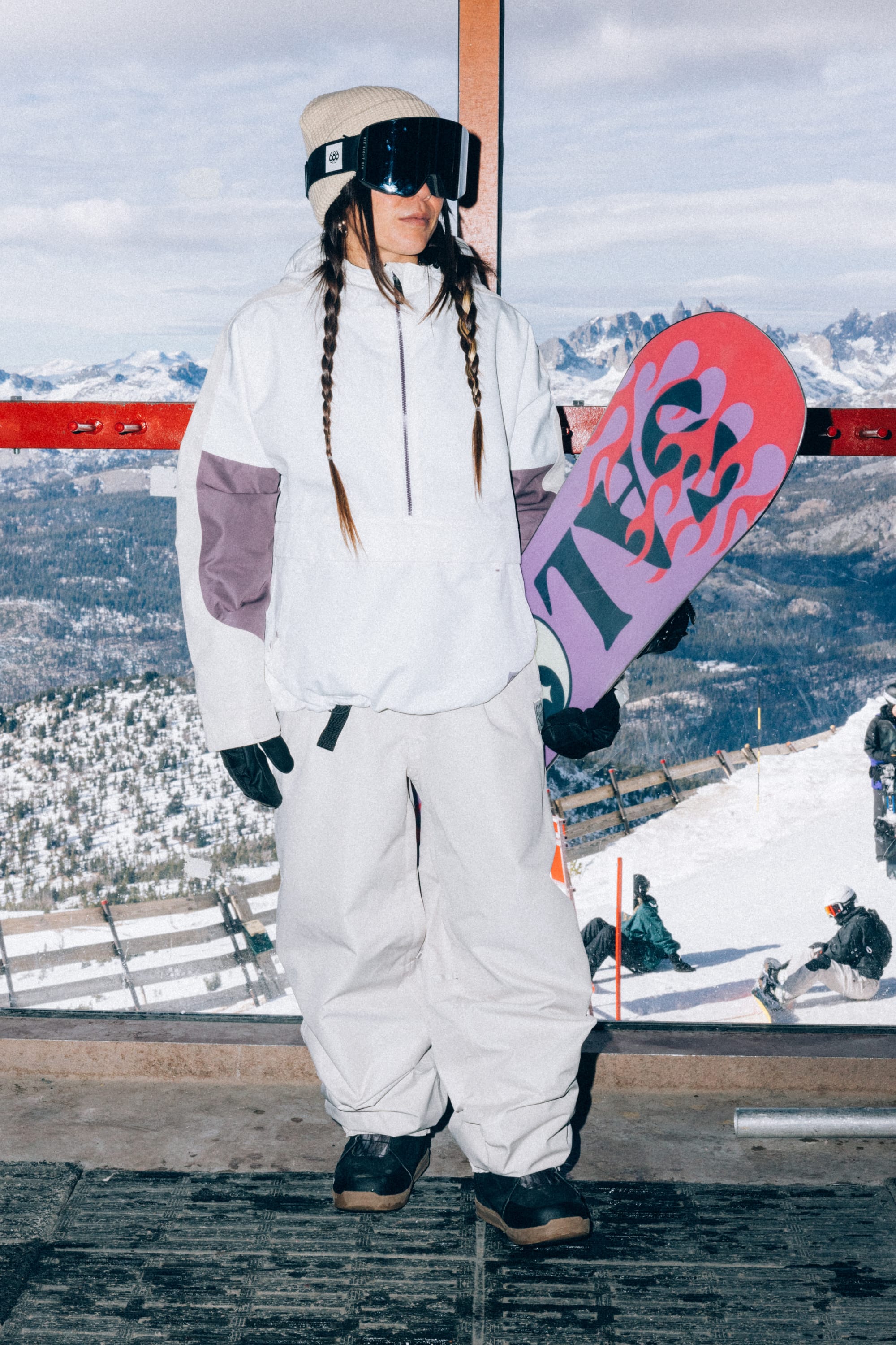 Wearing the 686 Women’s Outline™ Shell Pant, a snowboarder in white gear and goggles stands indoors holding a colorful snowboard, with snowy mountains and several people outside visible through the window behind her.