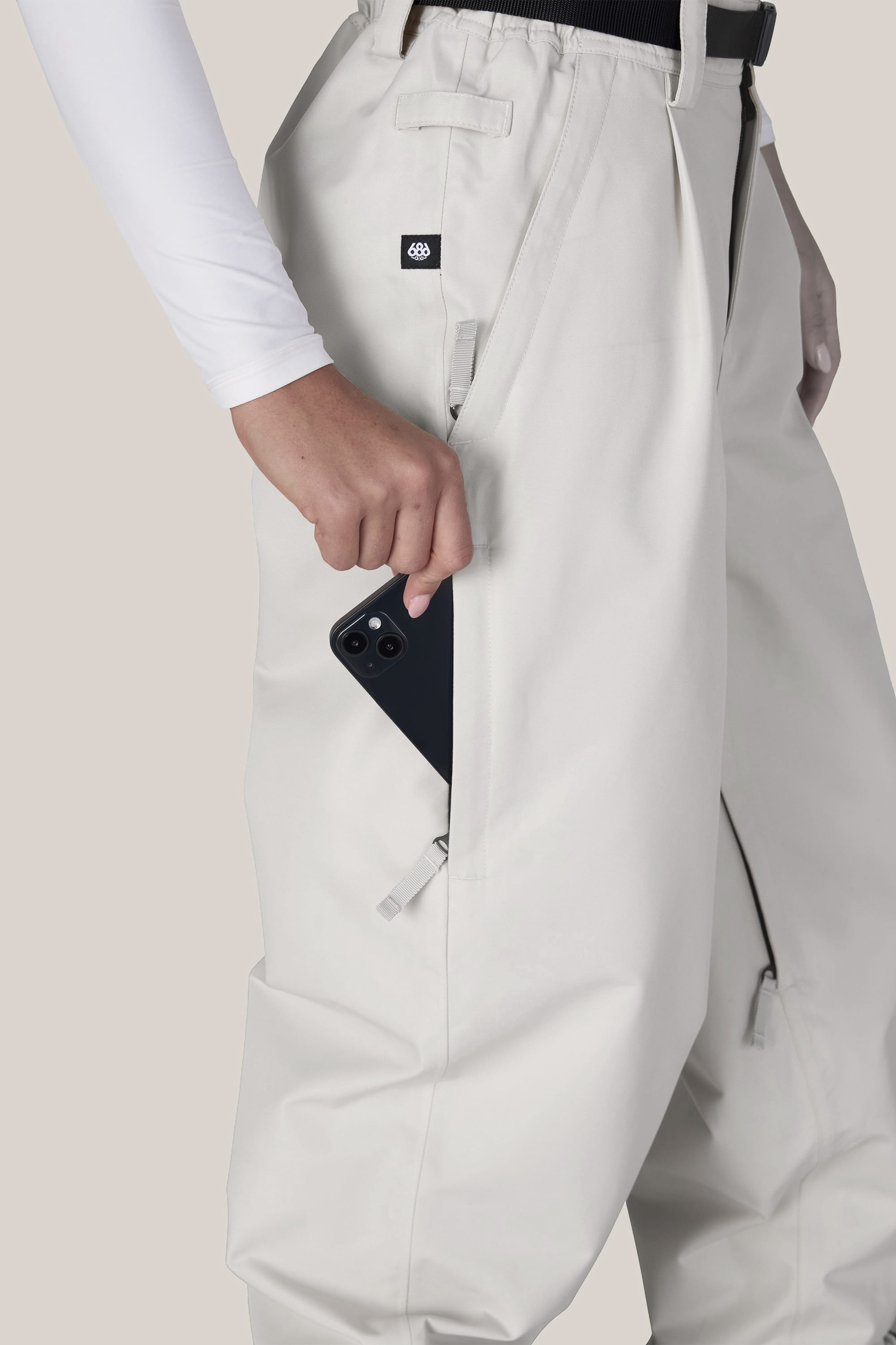 Wearing the 686 Women’s Outline™ Shell Pant, a person places a black smartphone into the zippered side pocket of their off-white pants.