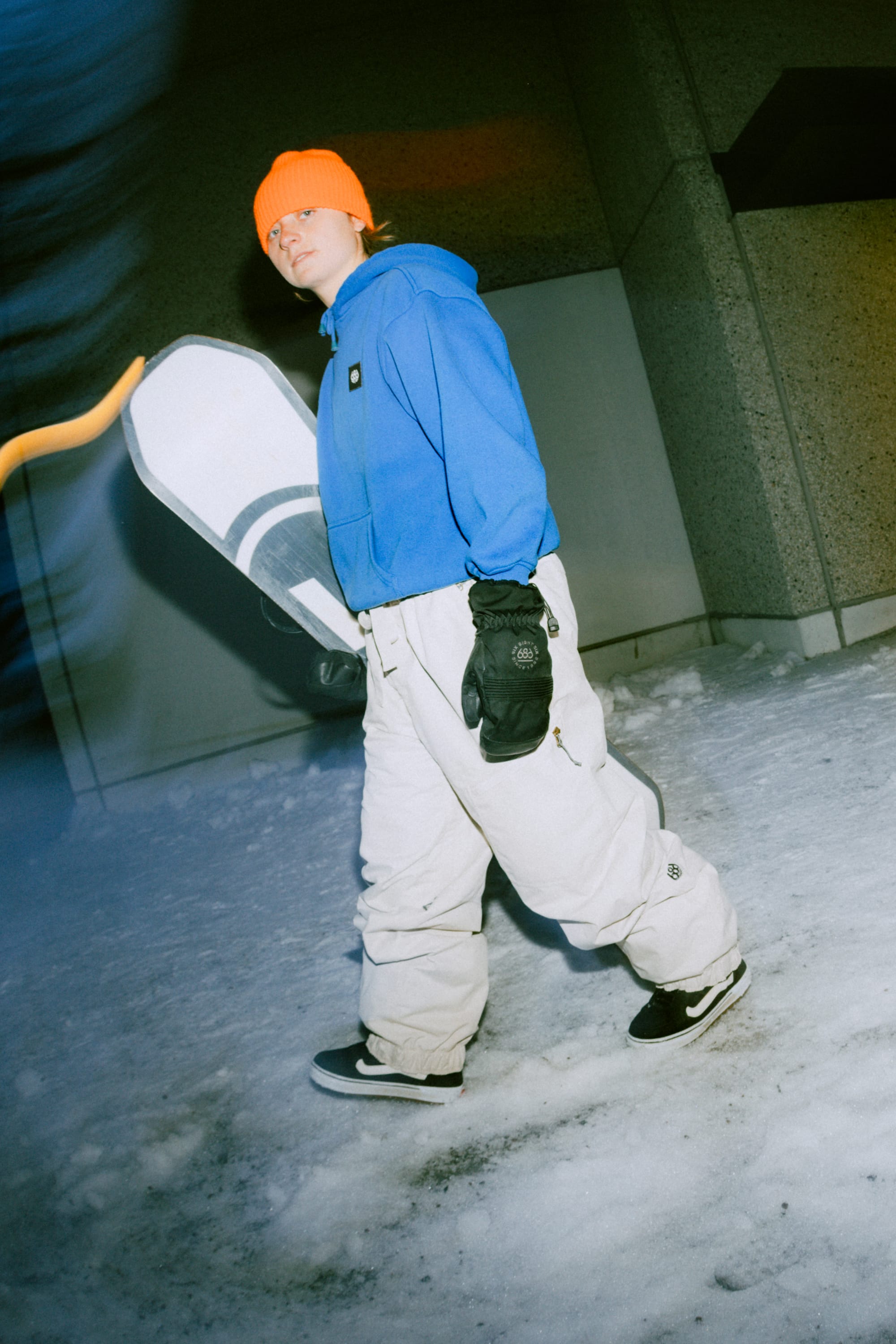A person in a bright orange beanie, blue hoodie, and 686 Women's Outline™ Shell Pant by 686 walks on snow at night, holding a snowboard and wearing black gloves and sneakers, with a building wall in the background.