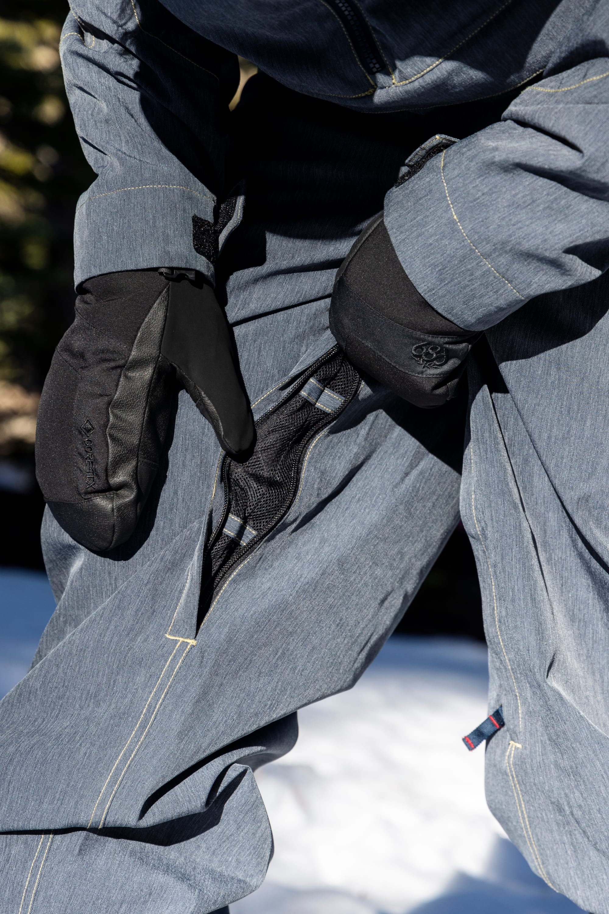 A person wearing the 686 Women’s Outline™ Shell Pant by 686 unzips a mesh vent on their pants while standing outdoors on snow, dressed in gray snow gear and black gloves.