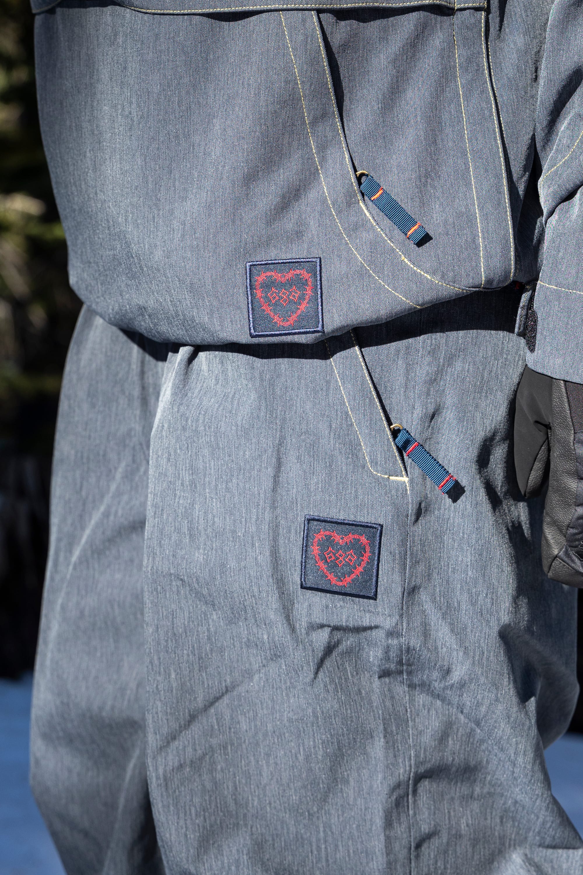 A close-up of a person outdoors wearing the 686 Women’s Outline™ Shell Pant, featuring gray fabric with blue and red heart patches. The person is also wearing a matching jacket and black glove, with trees and snow in the background.