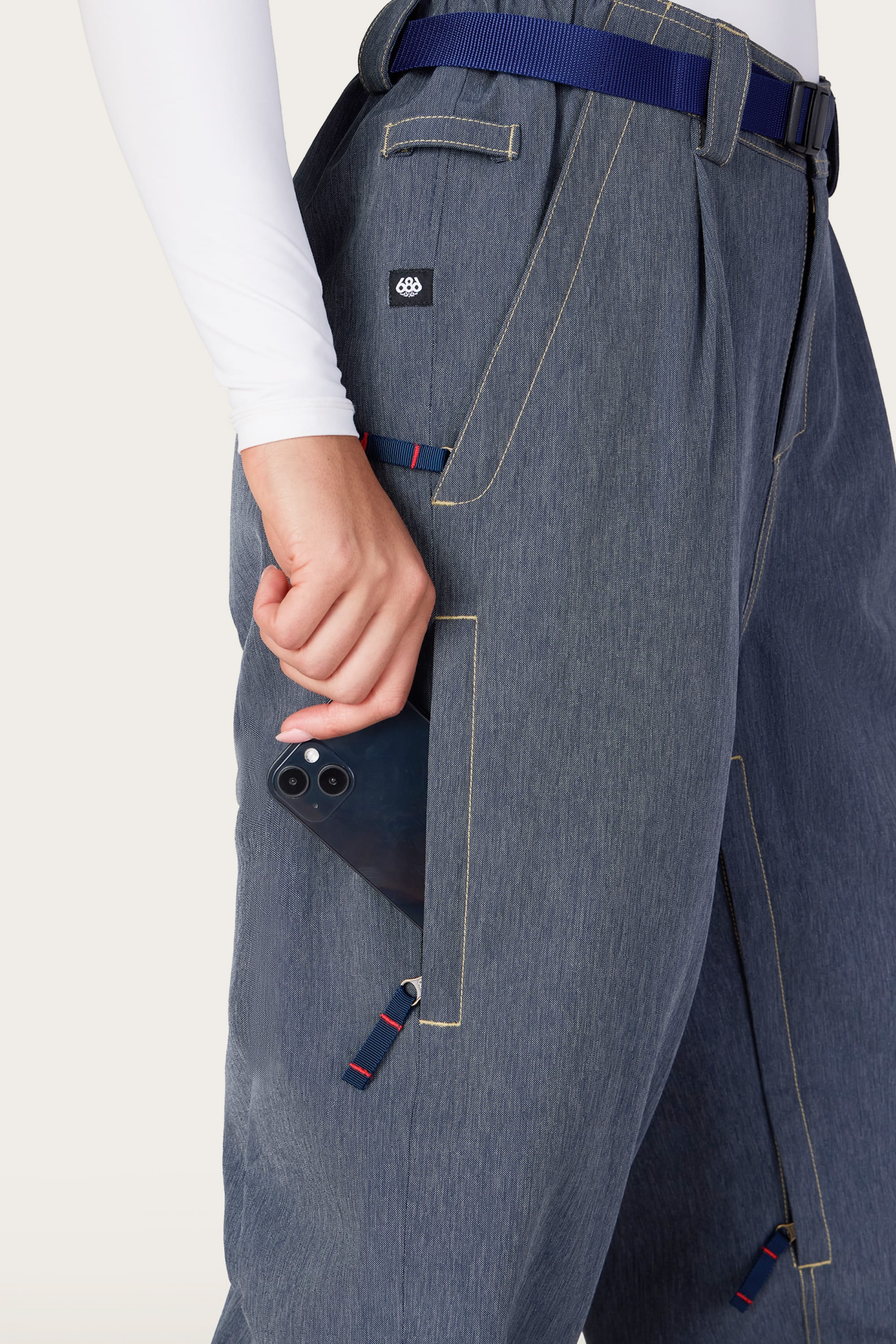 A person wearing the 686 Women’s Outline™ Shell Pant in blue-gray with white stitching puts a black smartphone into one of the large side pockets. The outfit features a blue belt and a long-sleeve white top, with only the lower torso and hand visible.