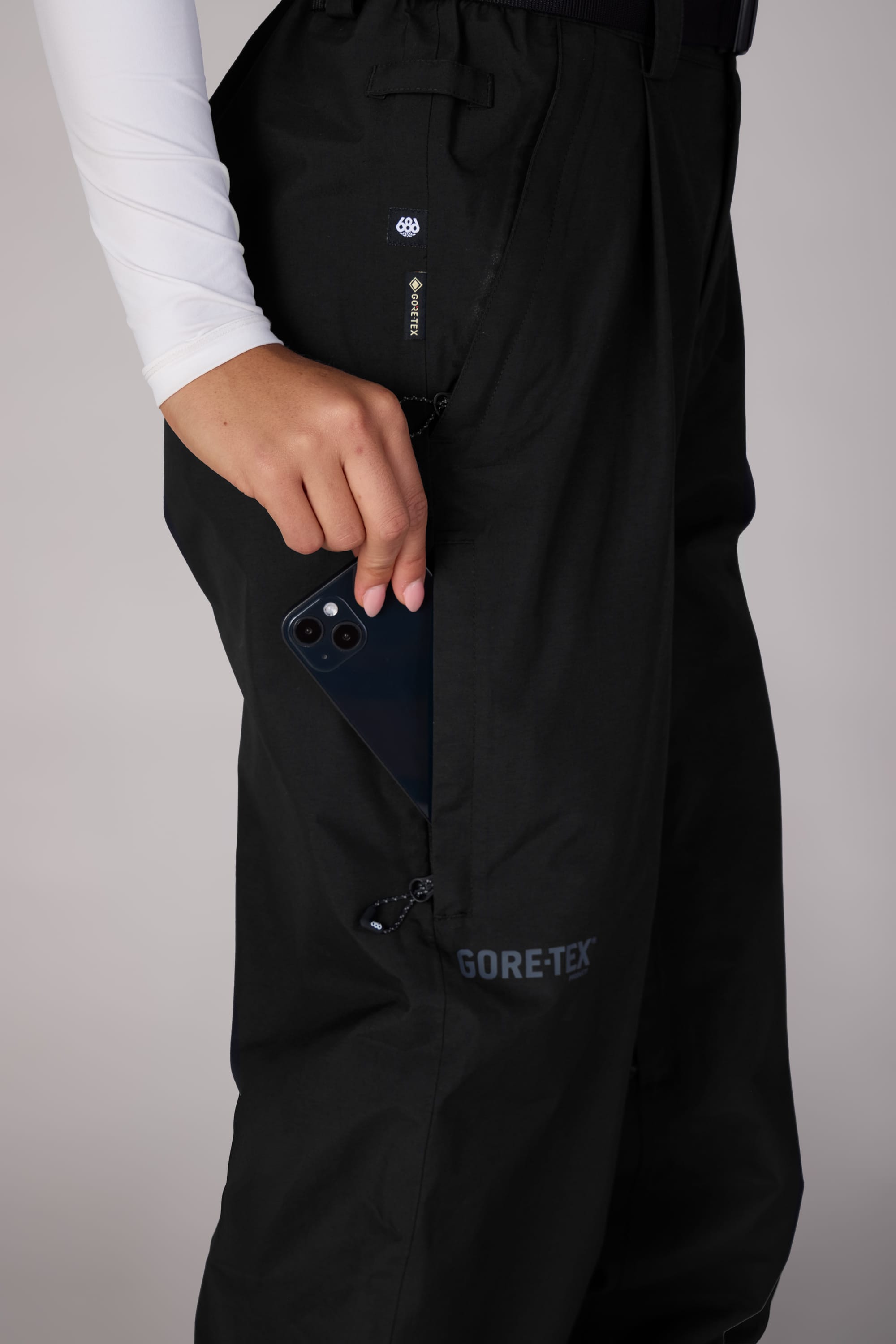 A woman wearing 686 Womens GORE-TEX Outline Shell Pants in black and a white long-sleeve top slips a black smartphone into the pants’ zippered side pocket.