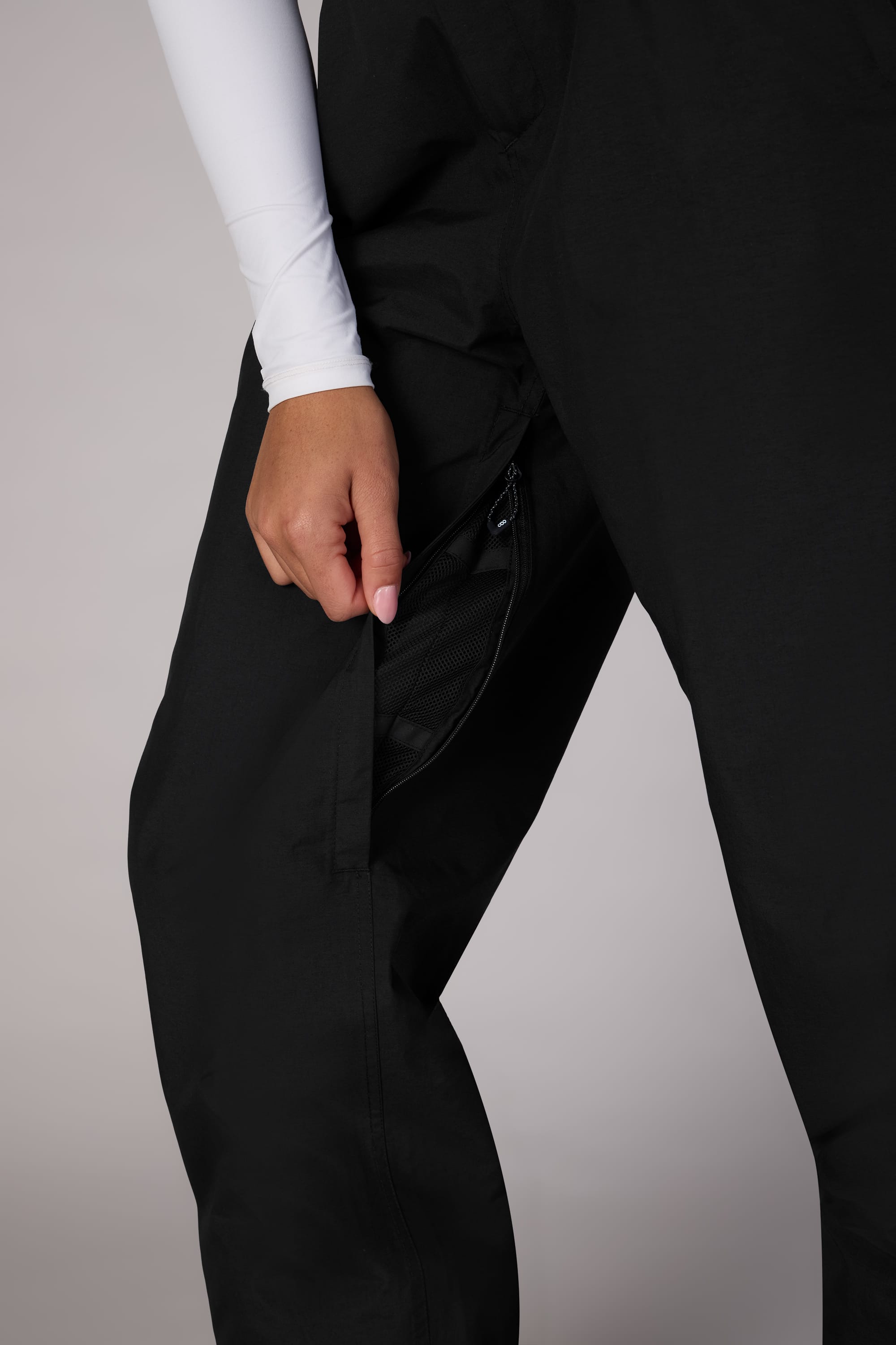 A person wearing black 686 Womens GORE-TEX Outline Shell Pants and a white long-sleeve shirt opens a zippered side pocket, with focus on the hand and pocket against a neutral gray background.
