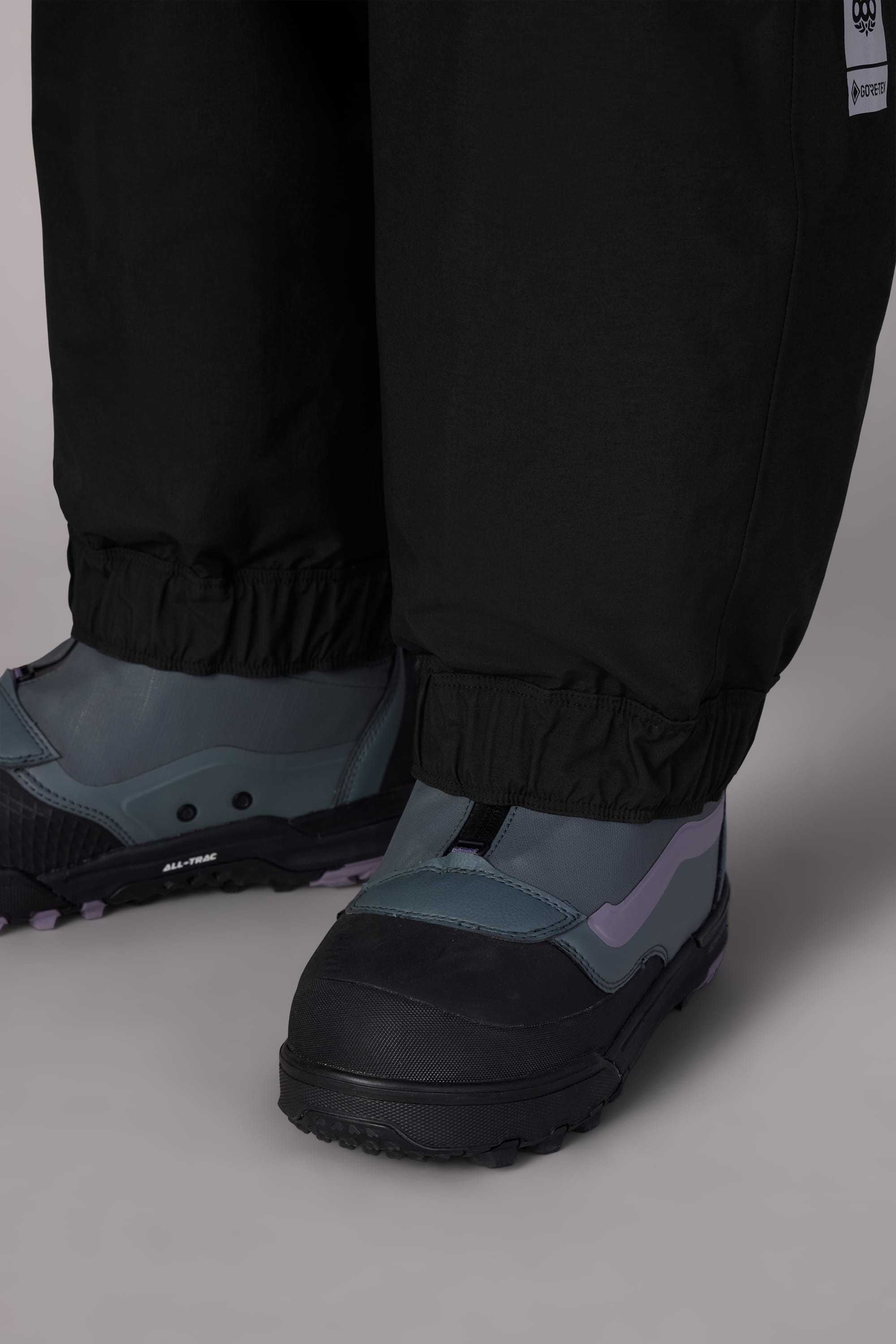 Close-up of a person wearing 686 Womens GORE-TEX Outline Shell Pant with elastic cuffs, paired with modern gray and black shoes featuring violet accents, standing on a light-colored surface.