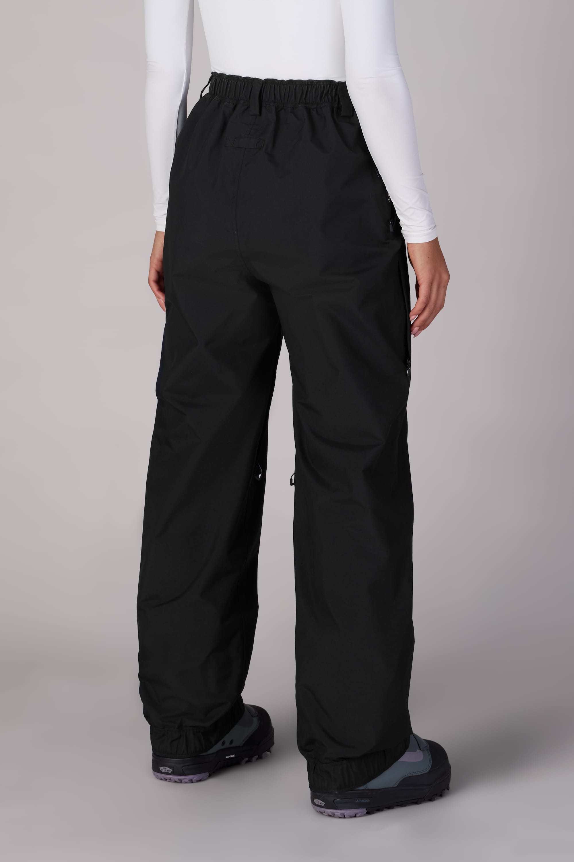 A person wearing 686 Womens GORE-TEX Outline Shell Pant, a fitted white long-sleeve top, and dark sneakers stands facing away against a light gray background.
