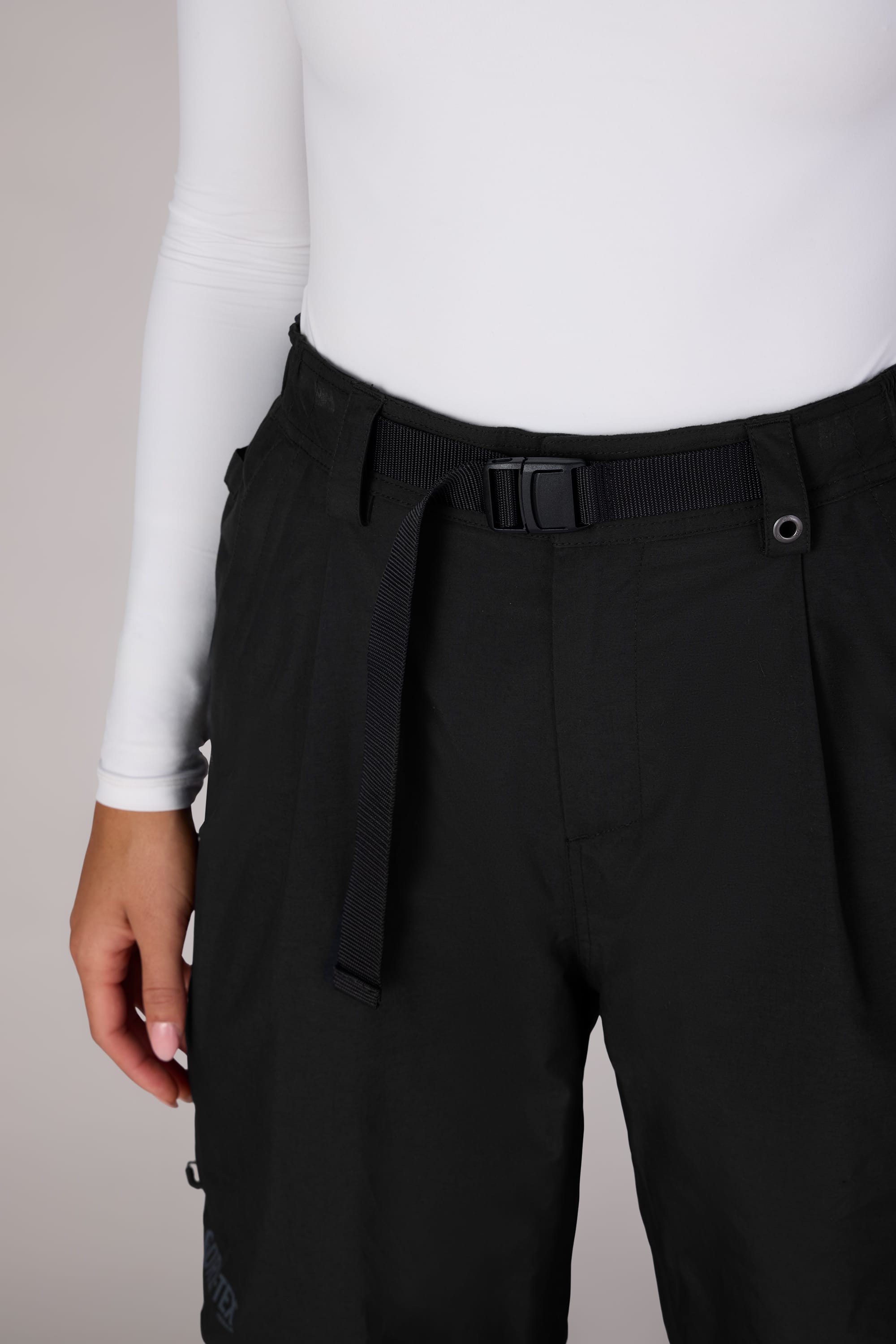 A person wears a white long-sleeve top and the 686 Womens GORE-TEX Outline Shell Pant in black, shown from waist to mid-thigh. Their left hand rests by their side.