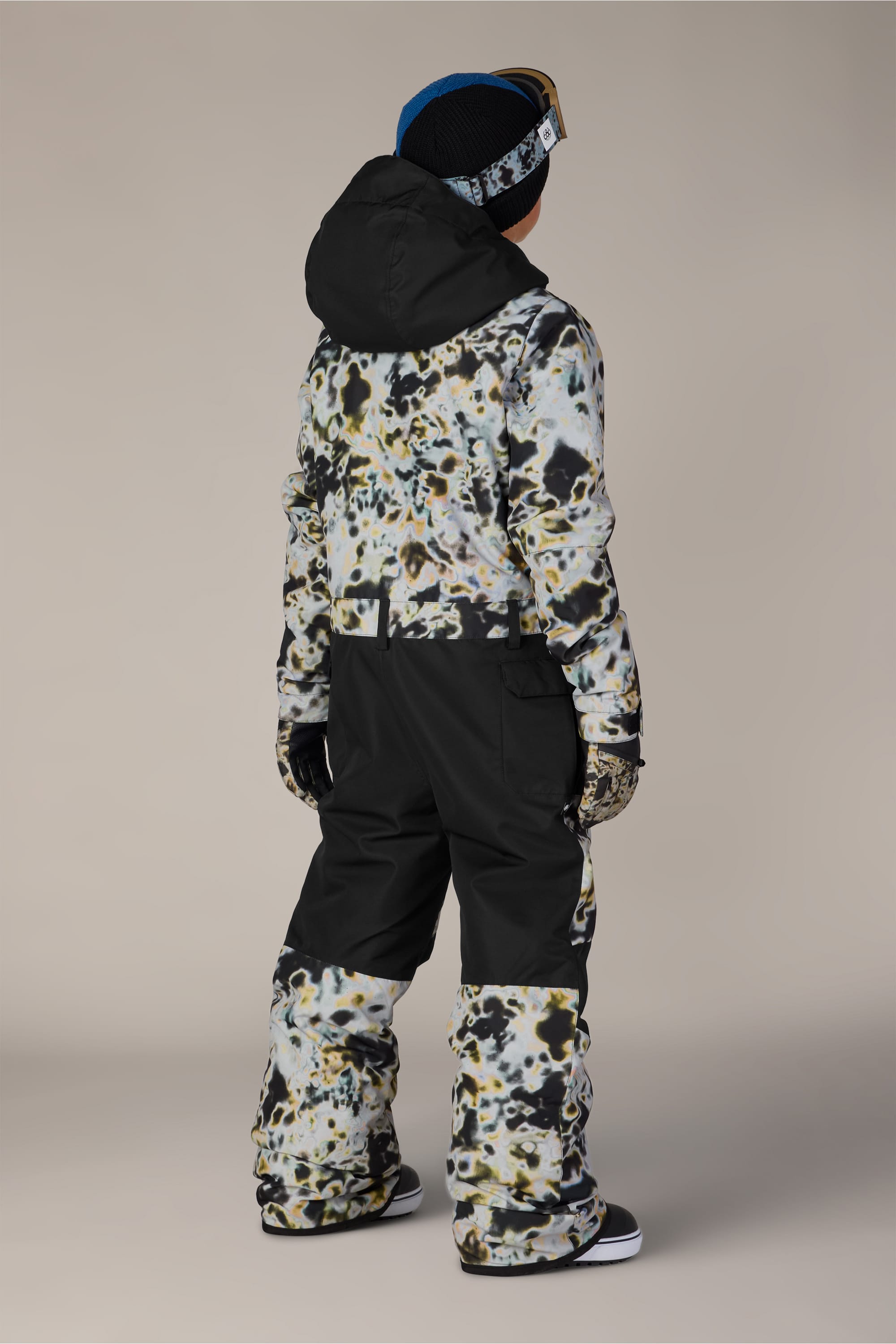 A child faces away, dressed in the 686 Boys’ Shazam Insulated One-Piece by 686, paired with black snow pants, gloves, a black hat, and blue ski goggles against a plain neutral background.
