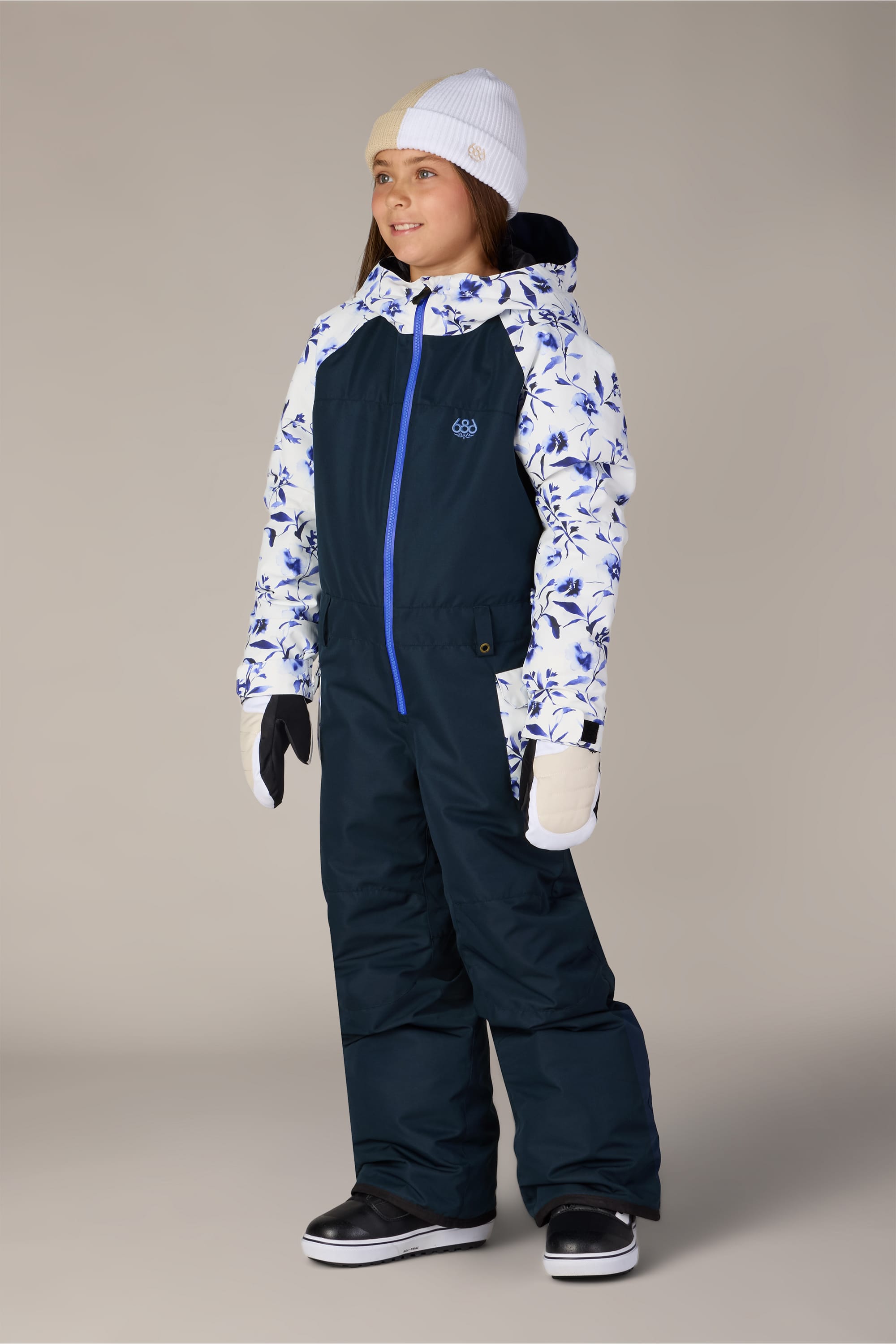 A girl wearing the 686 Girls’ Shine Insulated One-Piece by 686, along with a white beanie, mittens, and black boots, stands smiling against a plain background.