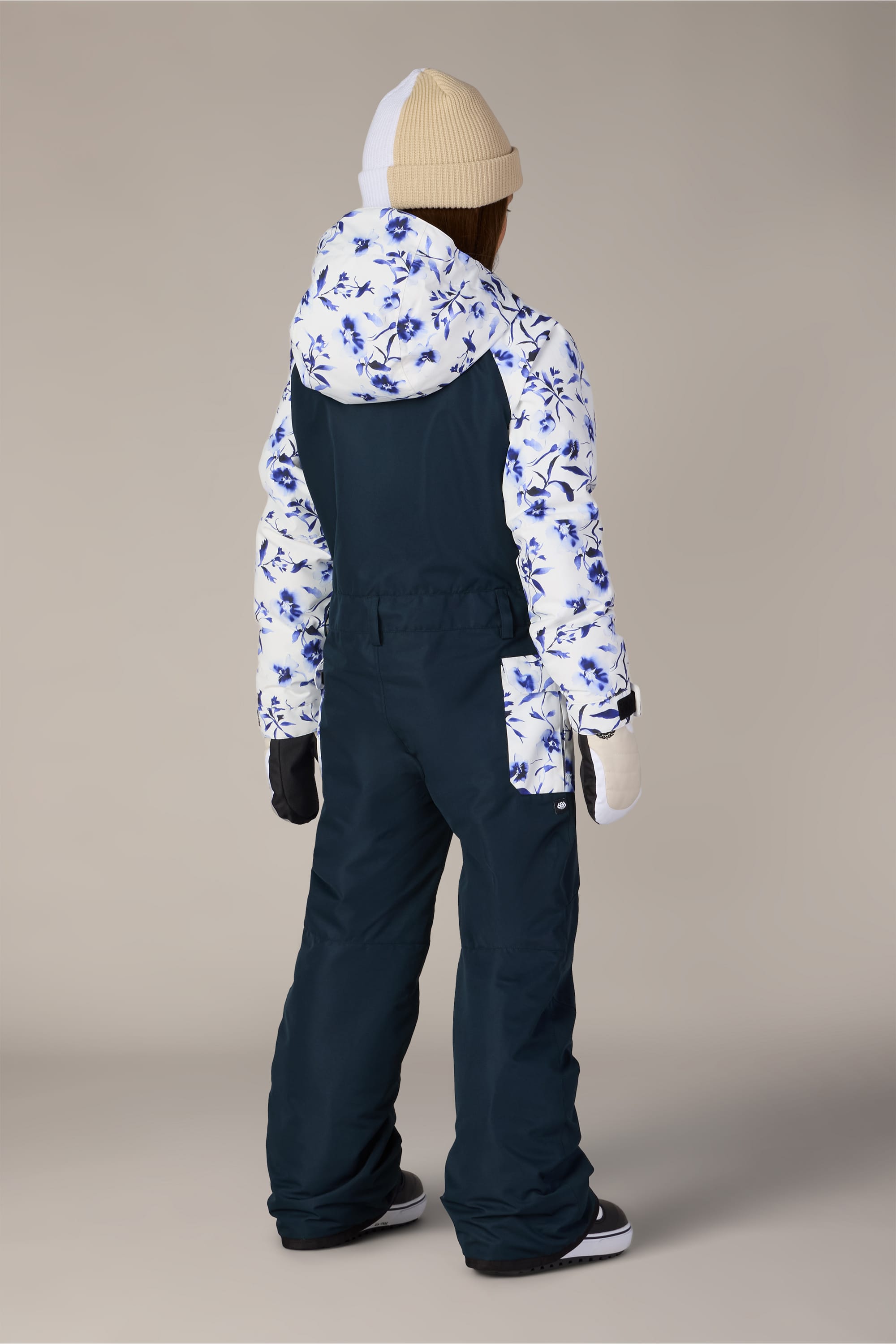 A person wearing the 686 Girls’ Shine Insulated One-Piece by 686, along with a beige beanie, mittens, and black boots, stands facing away on a plain background.