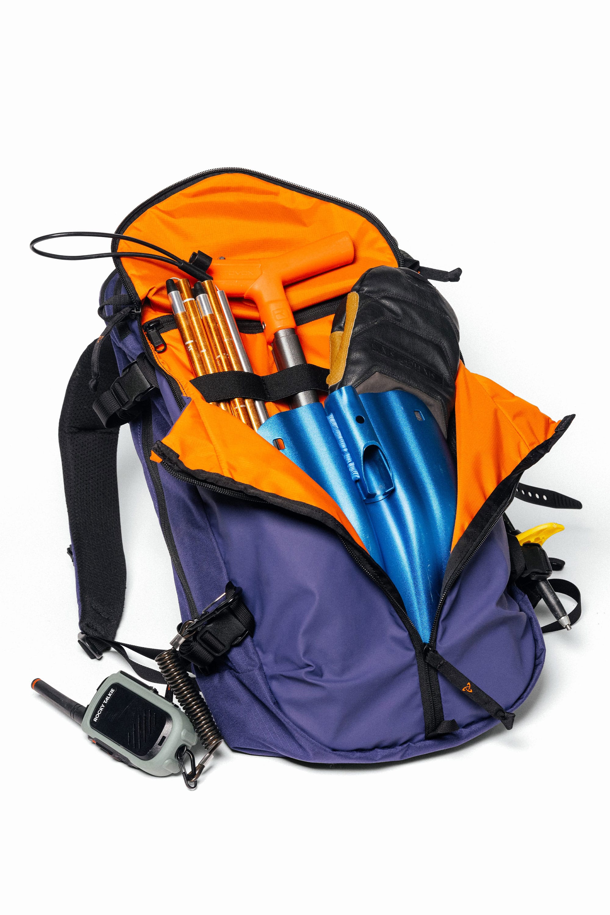The 686 Mystery Ranch x 686 Qi 22L Snow Pack is unzipped, revealing a blue shovel, an orange-handled tool, gold poles, and other gear inside. A walkie-talkie is attached to the outside of the pack.
