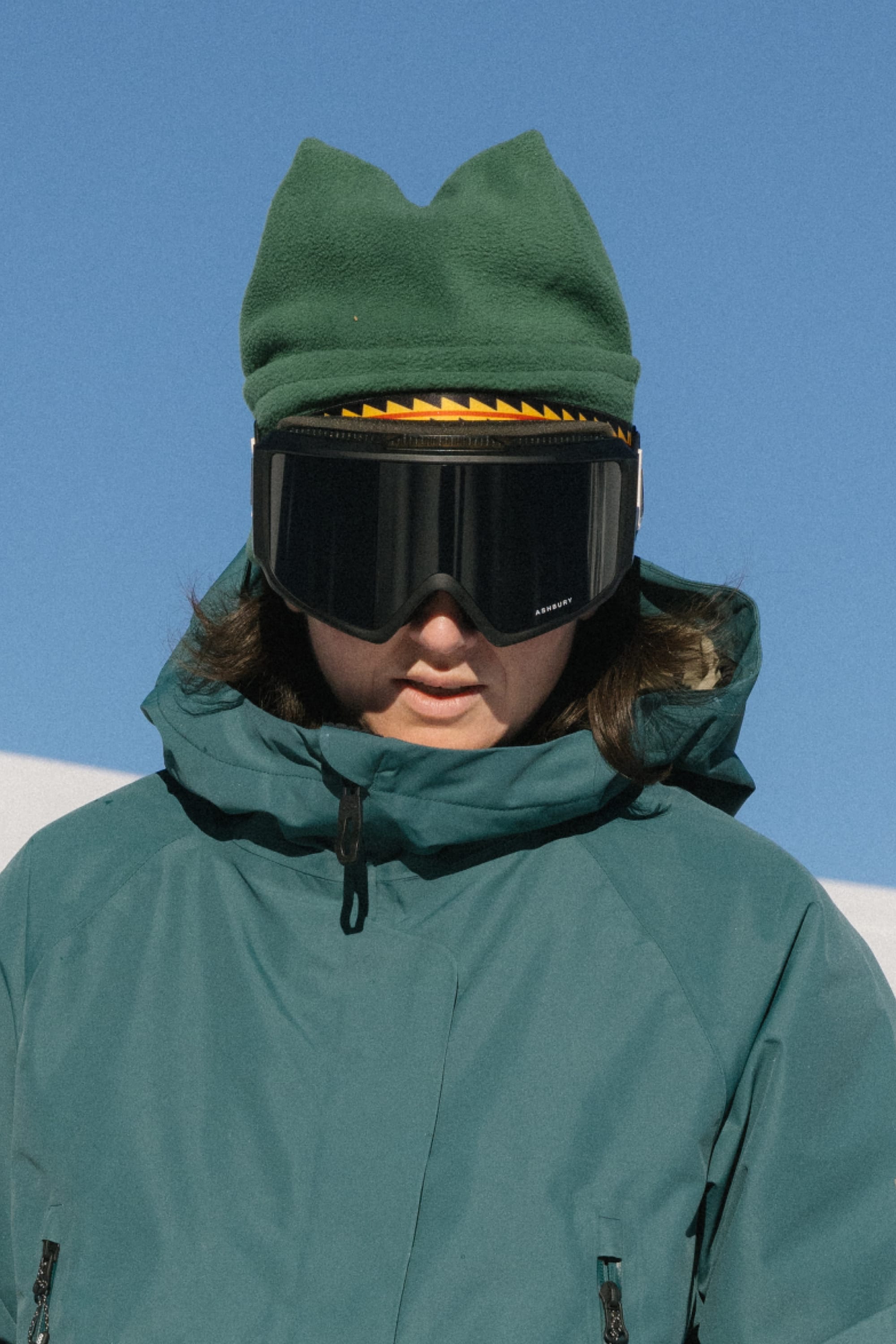 Wearing a green jacket, black ski goggles, and the 686 4 Points Fleece Hat by 686, a person stands outdoors on a sunny winter day beneath a clear blue sky.