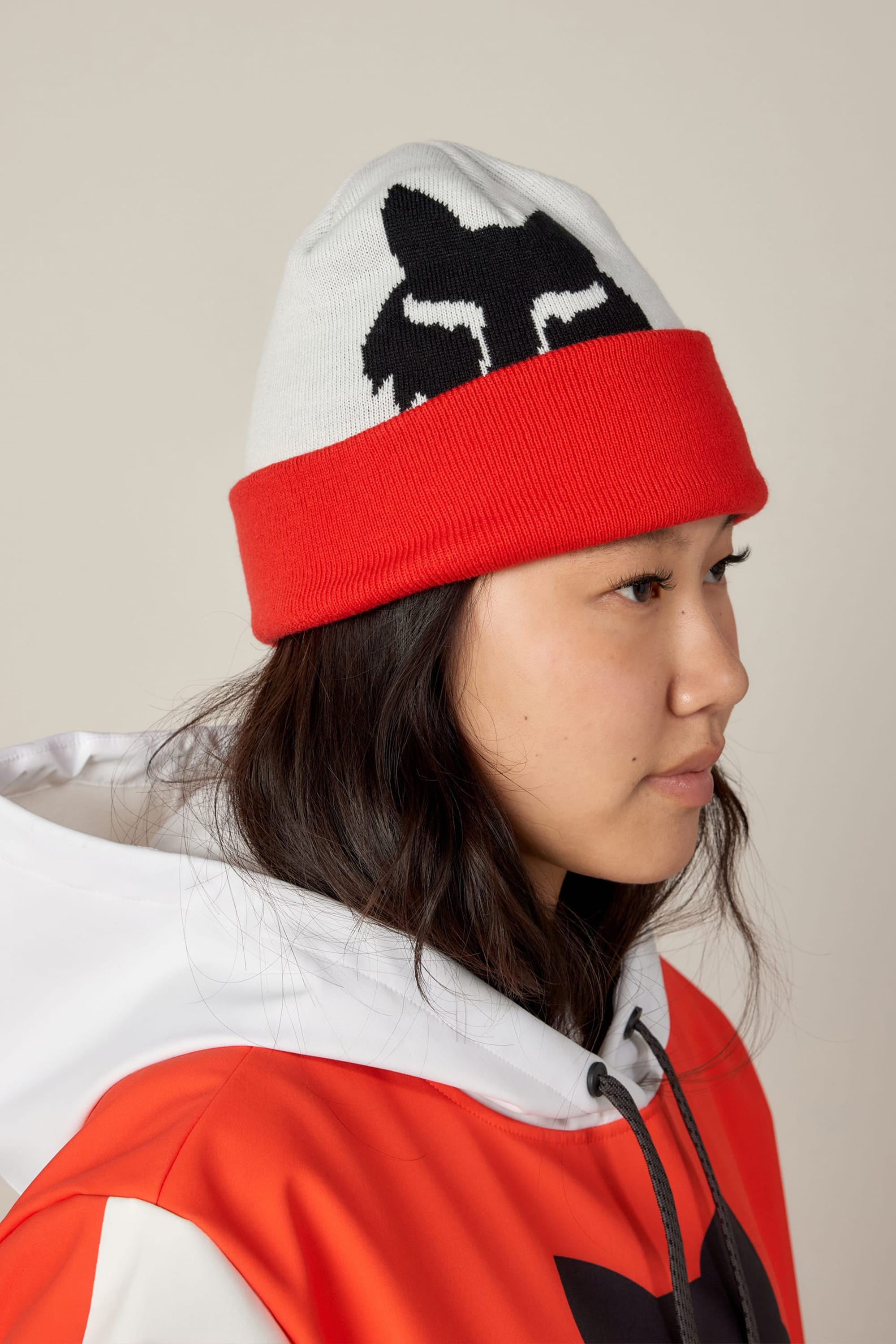 A person with long dark hair wears the 686 Fox Racing Cuff Beanie by 686, which is red and white with a black graphic design, along with a matching jacket. The person faces right against a plain background.