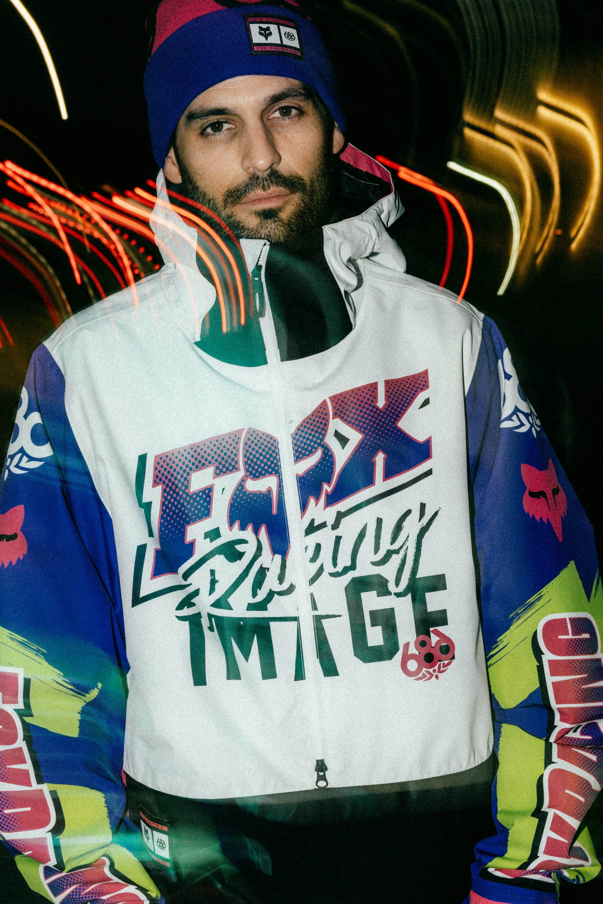 A man in a colorful jacket with bold graphics and FOX Racing IMAGE text stands against a dark background with light streaks, looking at the camera. He also wears a blue and pink 686 Fox Racing Cuff Beanie from 686.