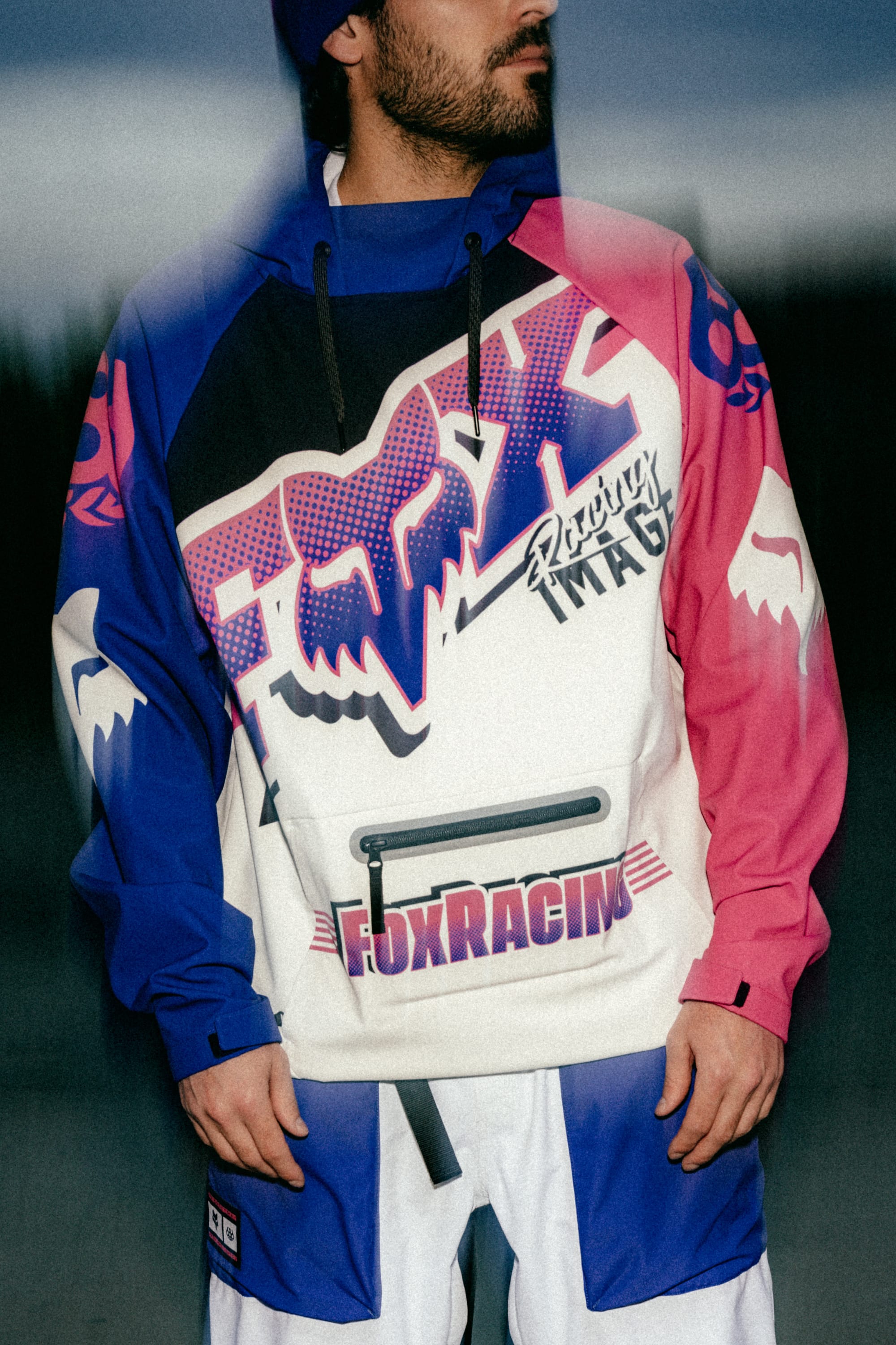 A person outdoors wears the 686 Men's Fox Racing Waterproof Hoody, featuring bold blue, pink, and white graphics with large FOX Racing text, stylized logos, and racing-themed details. The person's face is partially out of frame.