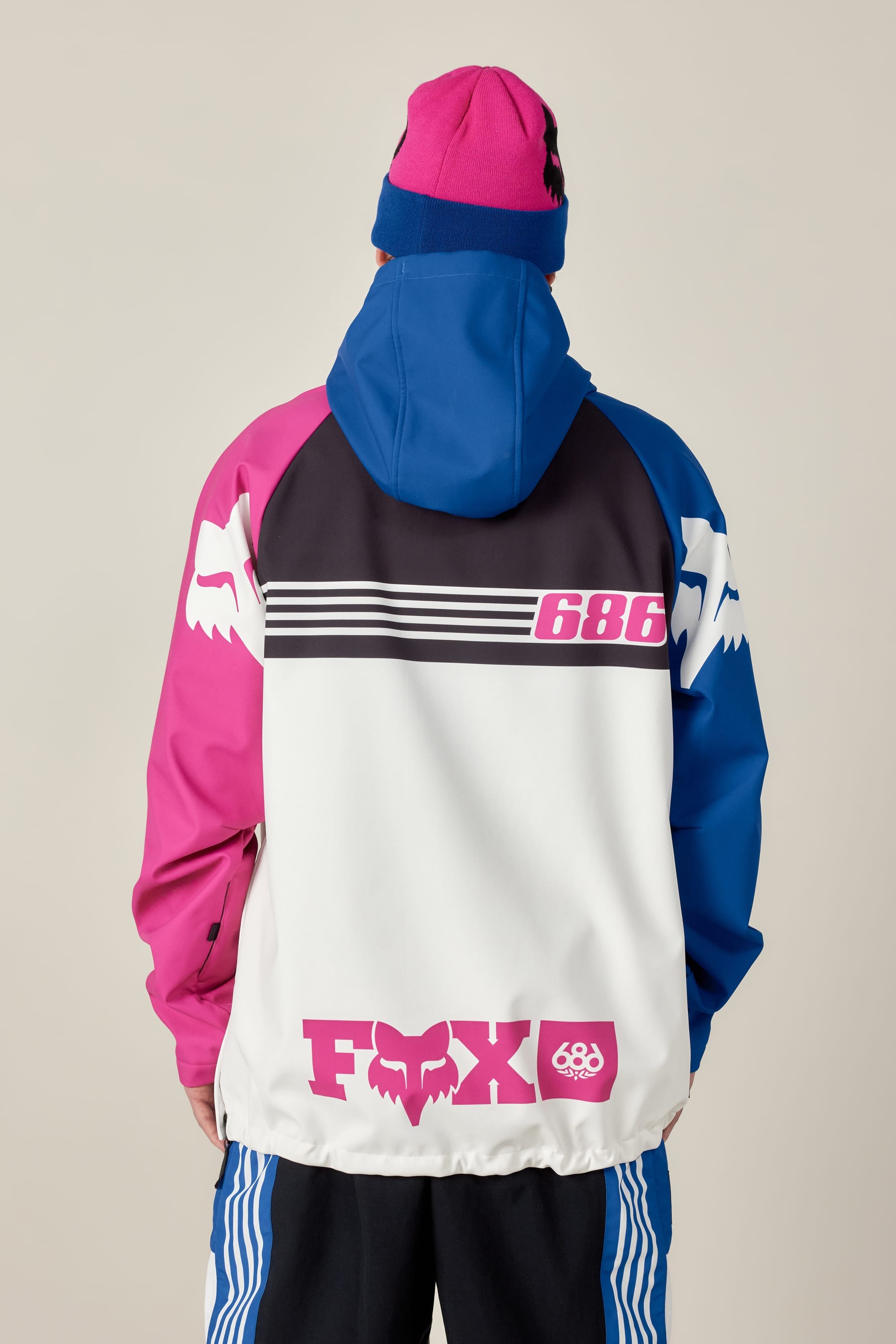 A person wearing the 686 Men's Fox Racing Waterproof Hoody with pink, blue, black, and white panels and FOX and 686 logos on the back, plus a matching pink and blue beanie, viewed from behind against a plain background.
