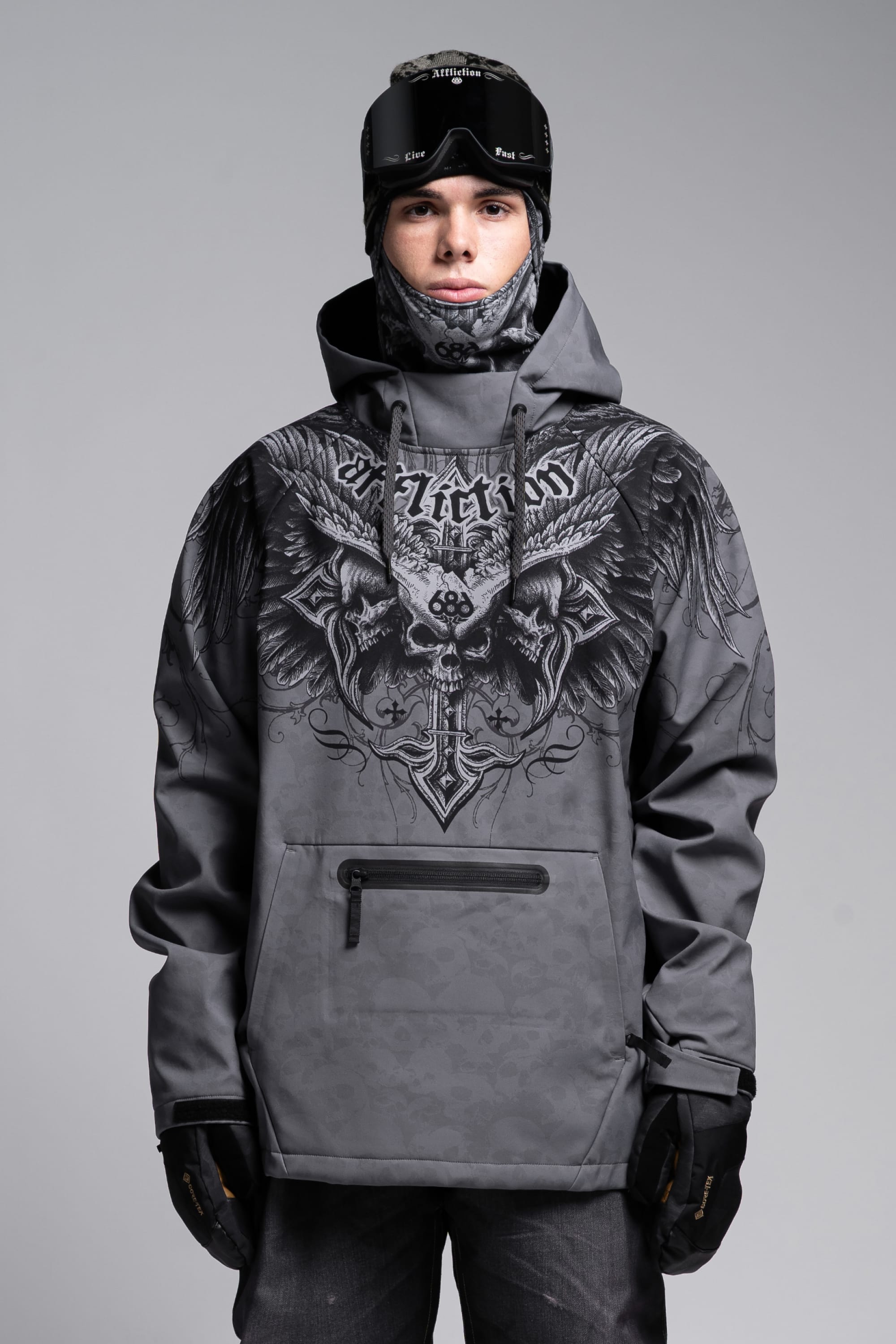 A person in a 686 Affliction Waterproof Hoody by 686, featuring an eagle and sword design, black gloves, a patterned neck warmer, and ski goggles on their forehead stands against a plain gray background.