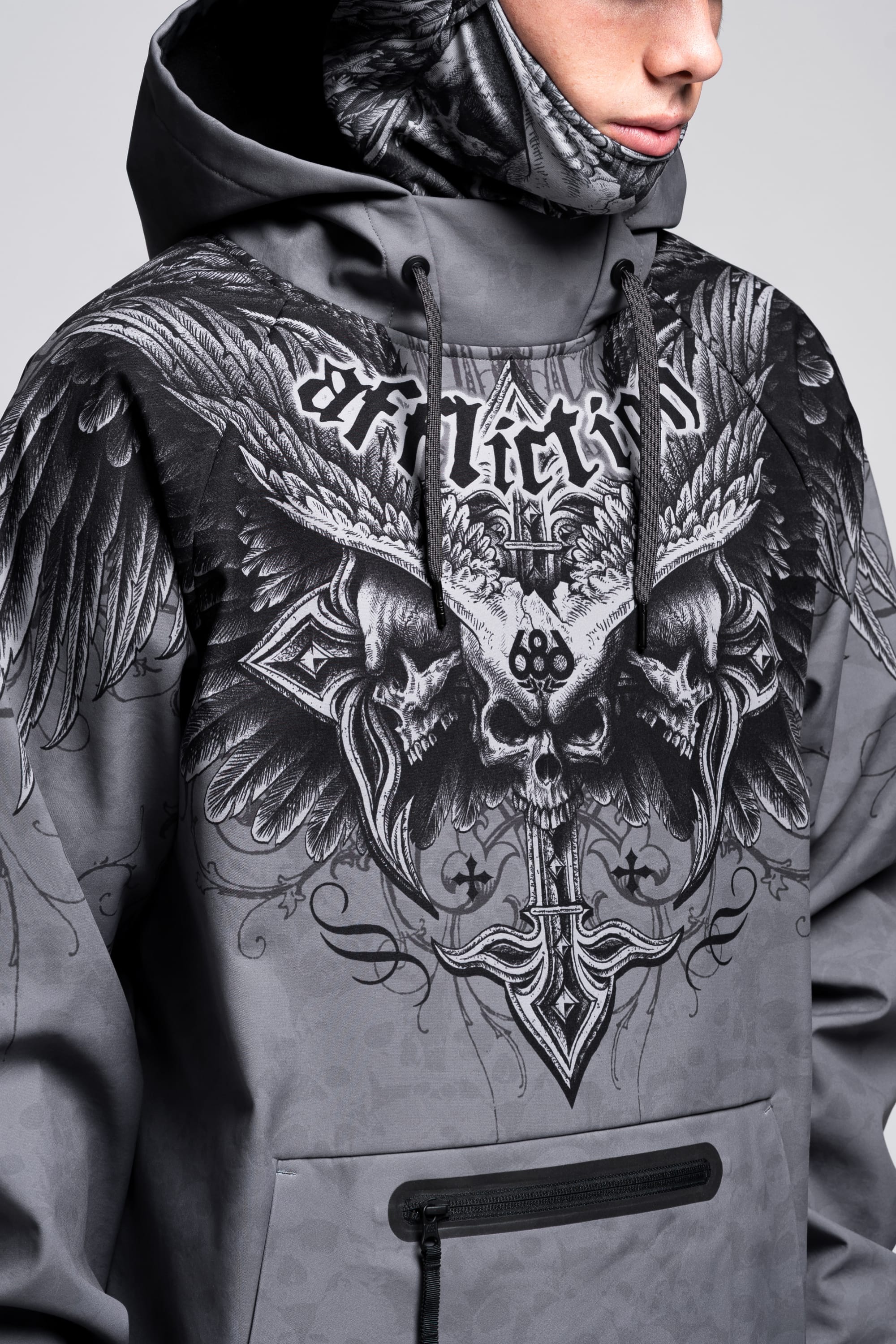 A person in a gray 686 Affliction Waterproof Hoody, featuring bold winged skull graphics, ornate designs, and "Affliction" across the chest; only the lower half of their face is visible.
