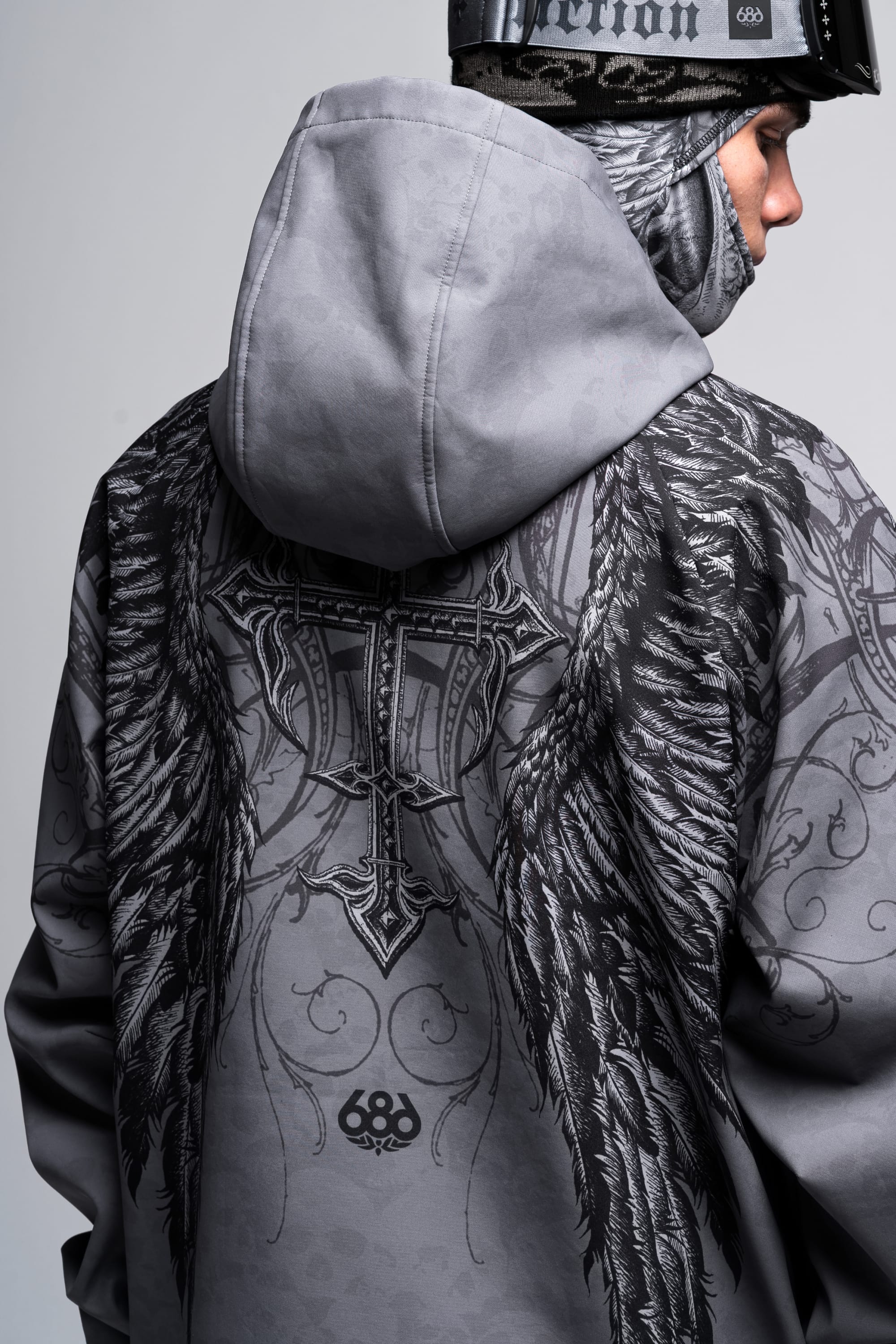 A person wearing the 686 Affliction Waterproof Hoody by 686, featuring black wing and cross designs on the back, along with a helmet and face covering, viewed from behind and slightly to the side.