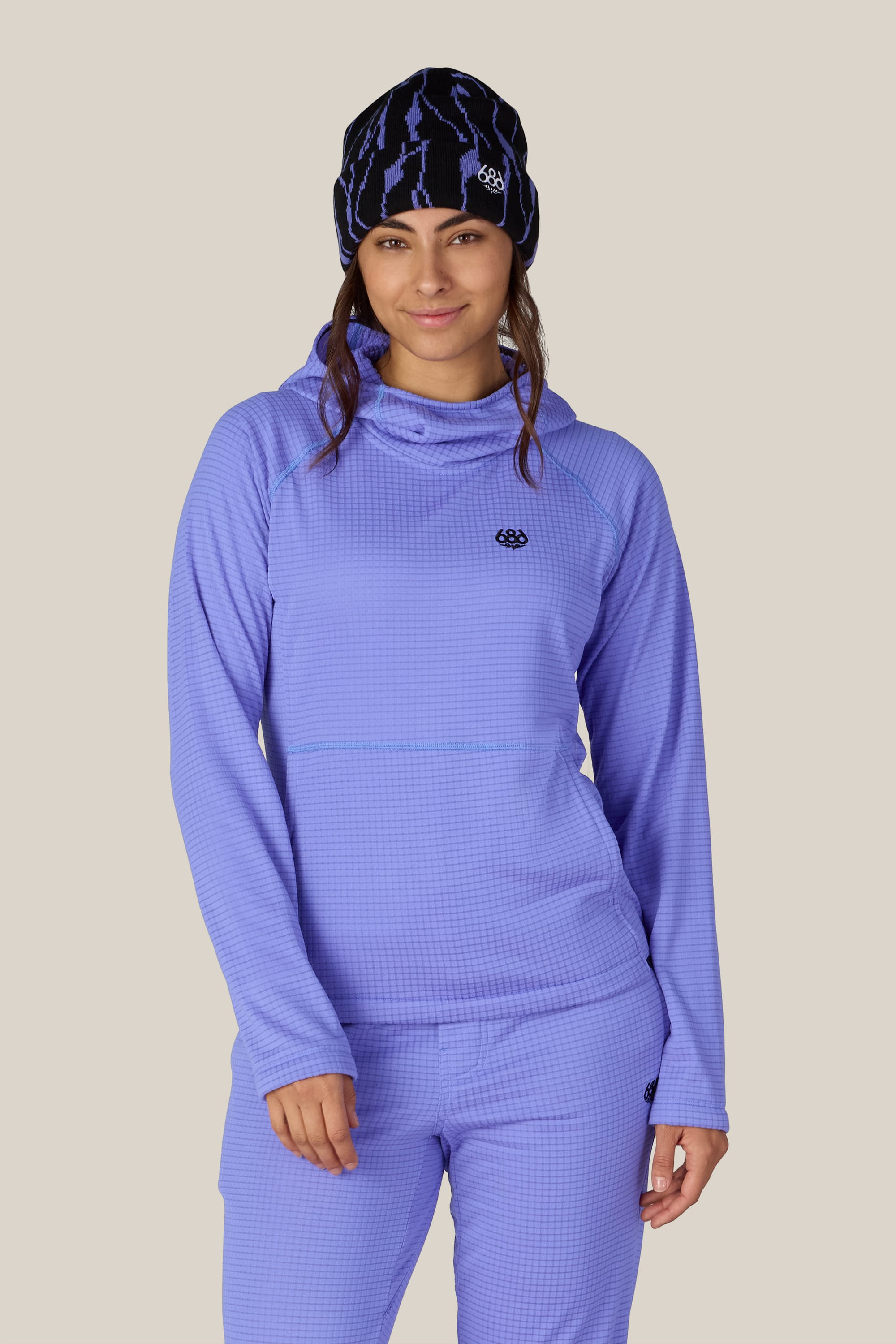 A woman stands against a plain beige background, smiling slightly with her hand by her side, wearing the 686 Women’s Mission Grid™ Hoody in light purple polyester knit paired with matching pants, and accessorized with a black and blue patterned beanie.