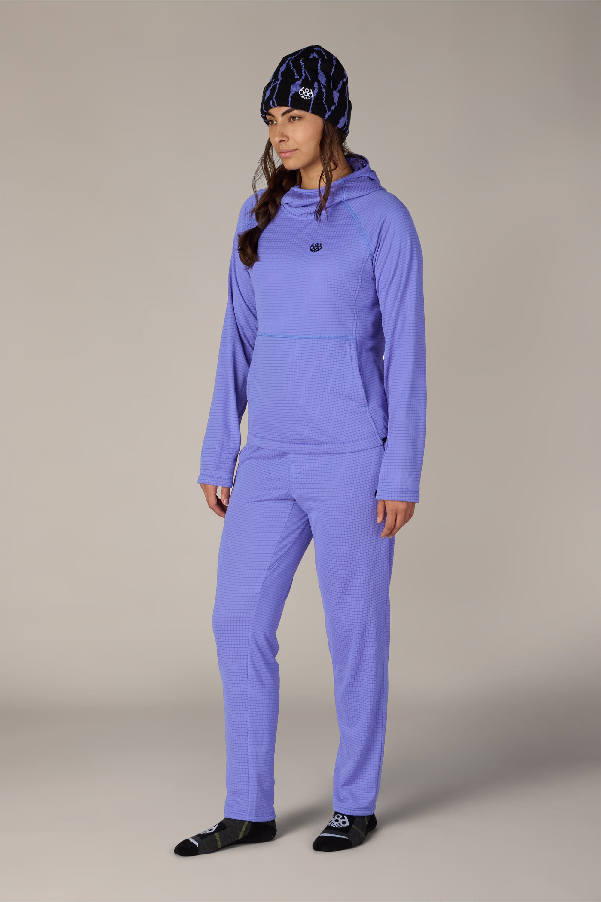 A person stands against a plain light background, looking slightly to the side, dressed in a matching light purple set and a patterned beanie with black socks. The cozy ensemble calls to mind the 686 Women’s Mission Grid™ Hoody by 686—soft, comfortable, and ideal for casual style.