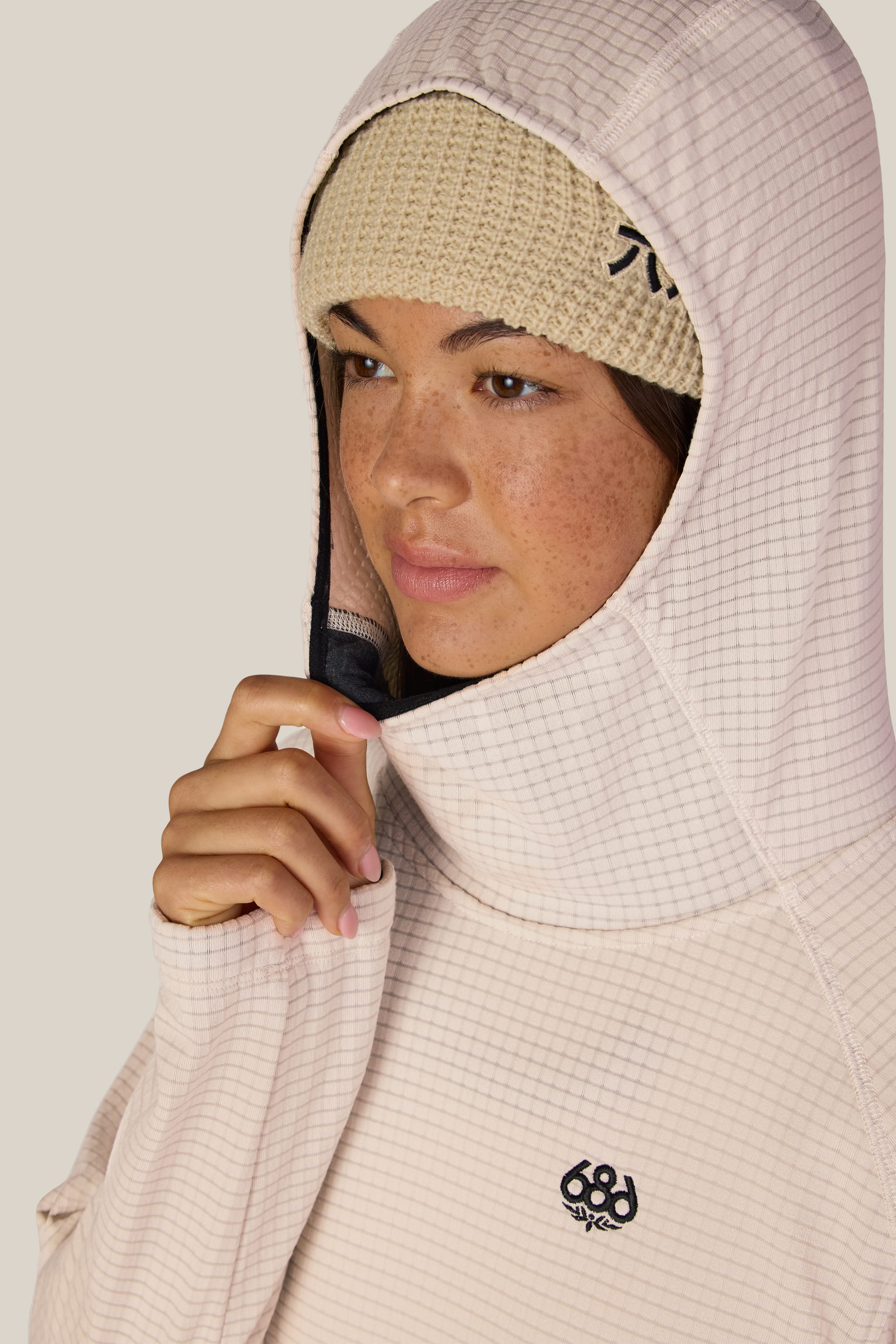 A woman wearing a beige knit beanie and the 686 Women’s Mission Grid™ Hoody gently holds the hood near her face, looking to the side with a neutral expression.