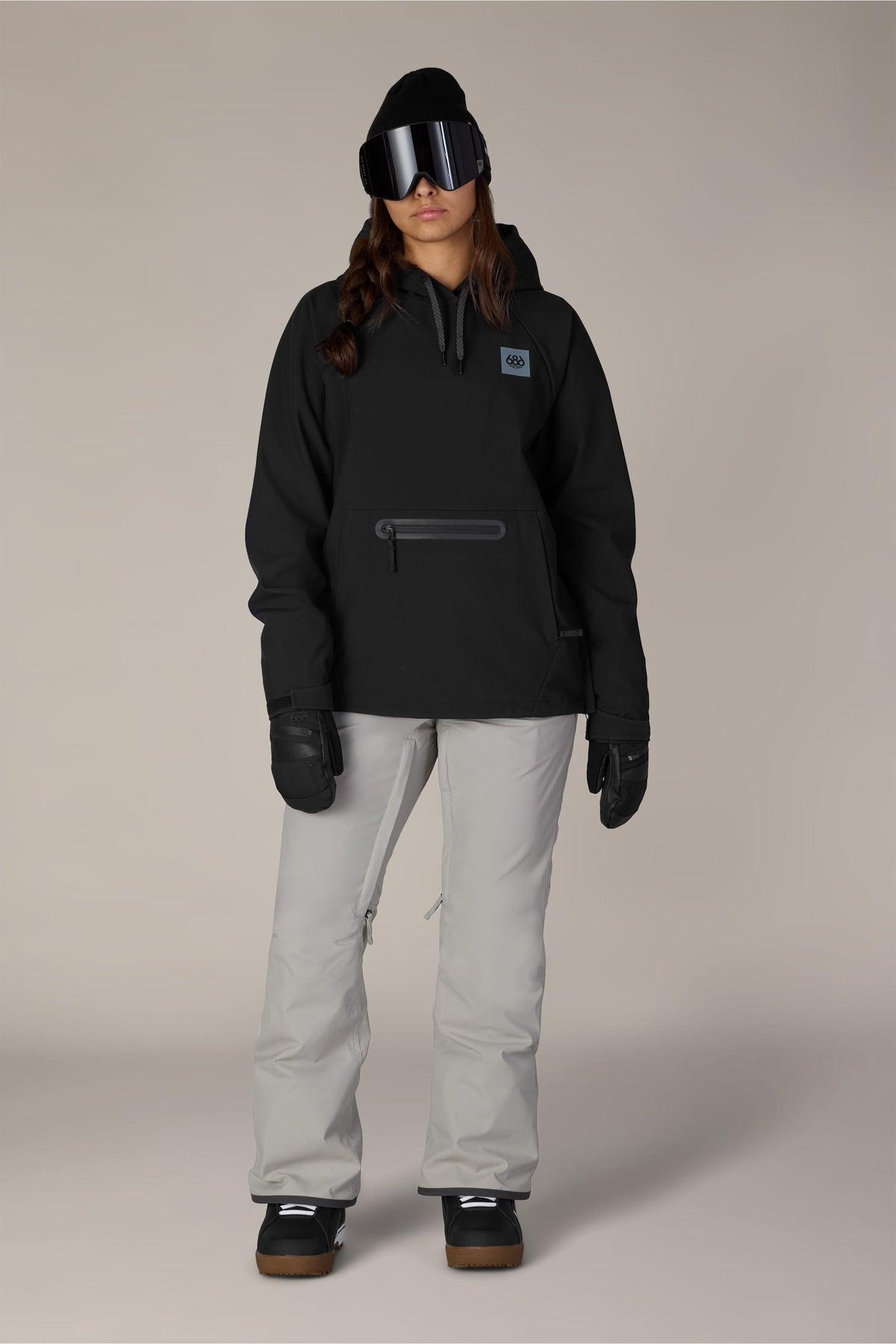 A person wearing the 686 Women’s Waterproof Hoody by 686, paired with light gray snow pants, black gloves, a black beanie, and large ski goggles stands facing forward against a plain background.