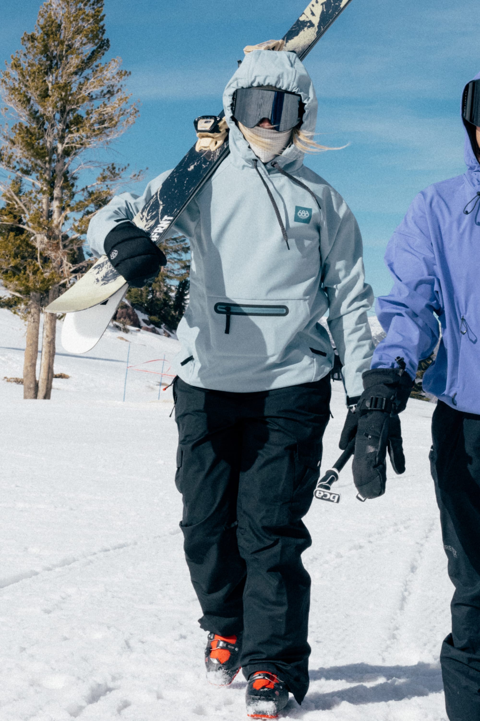 Wearing the 686 Women’s Waterproof Hoody, a woman in winter gear and goggles carries her skis on her shoulder, walking through snowy terrain alongside another similarly dressed companion.