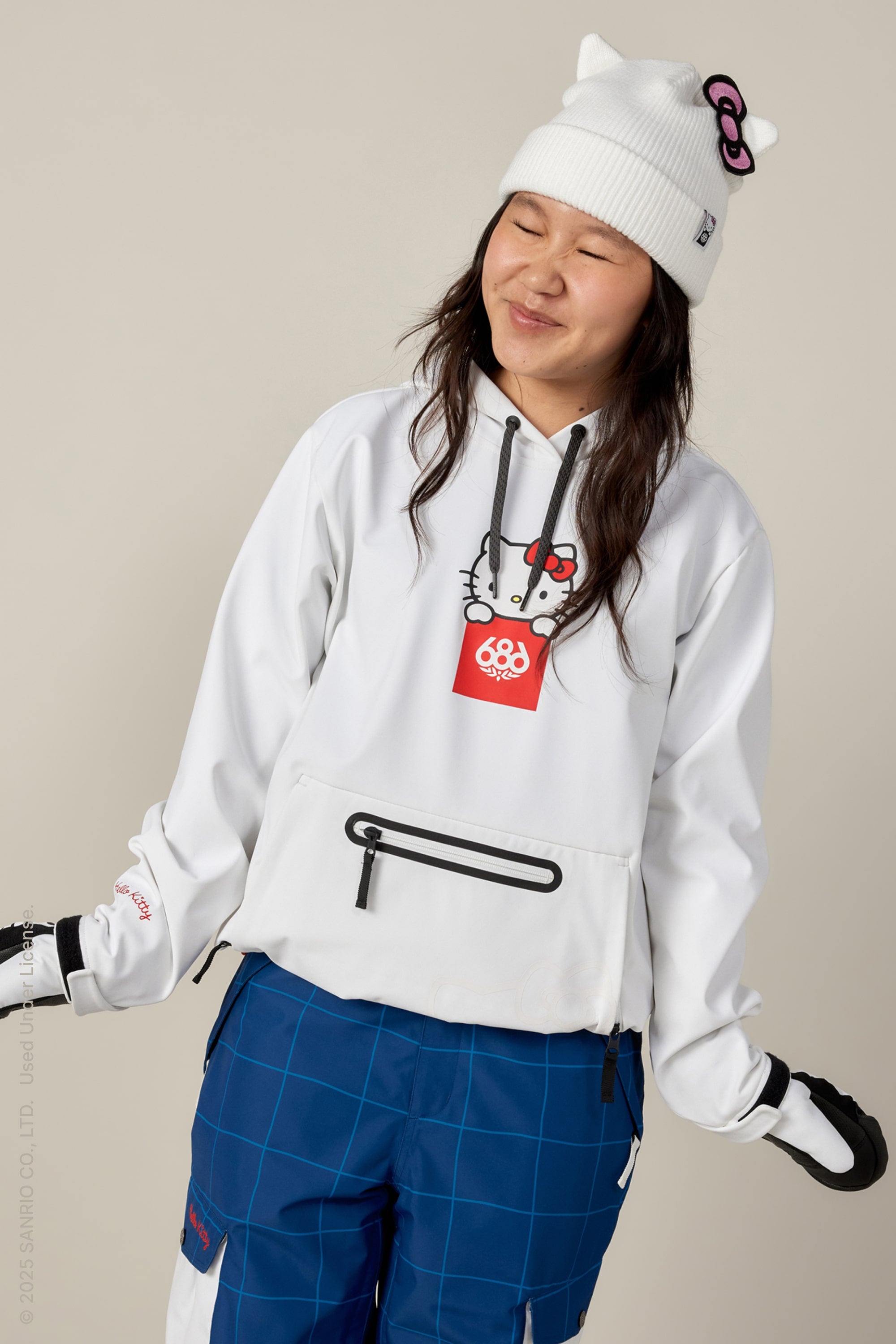 A person smiles with eyes closed against a plain light background, wearing a white Hello Kitty beanie, 686 Women's Waterproof Hoody by 686 featuring Hello Kitty graphics, blue grid-patterned pants, and black-and-white gloves.