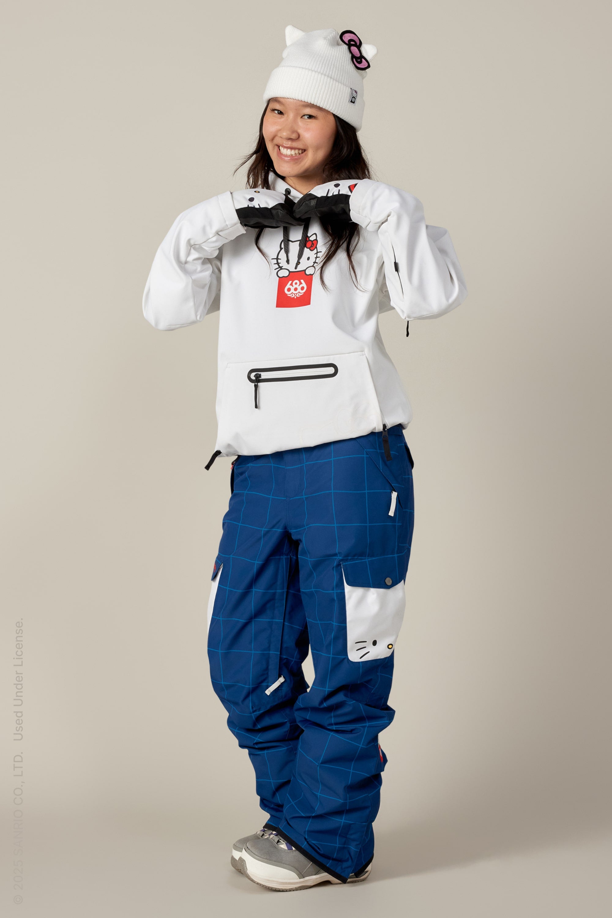 A smiling person poses against a plain background wearing the 686 Women's Waterproof Hoody by 686 along with blue checkered Hello Kitty pants and a white Hello Kitty beanie, hands up near their chest.