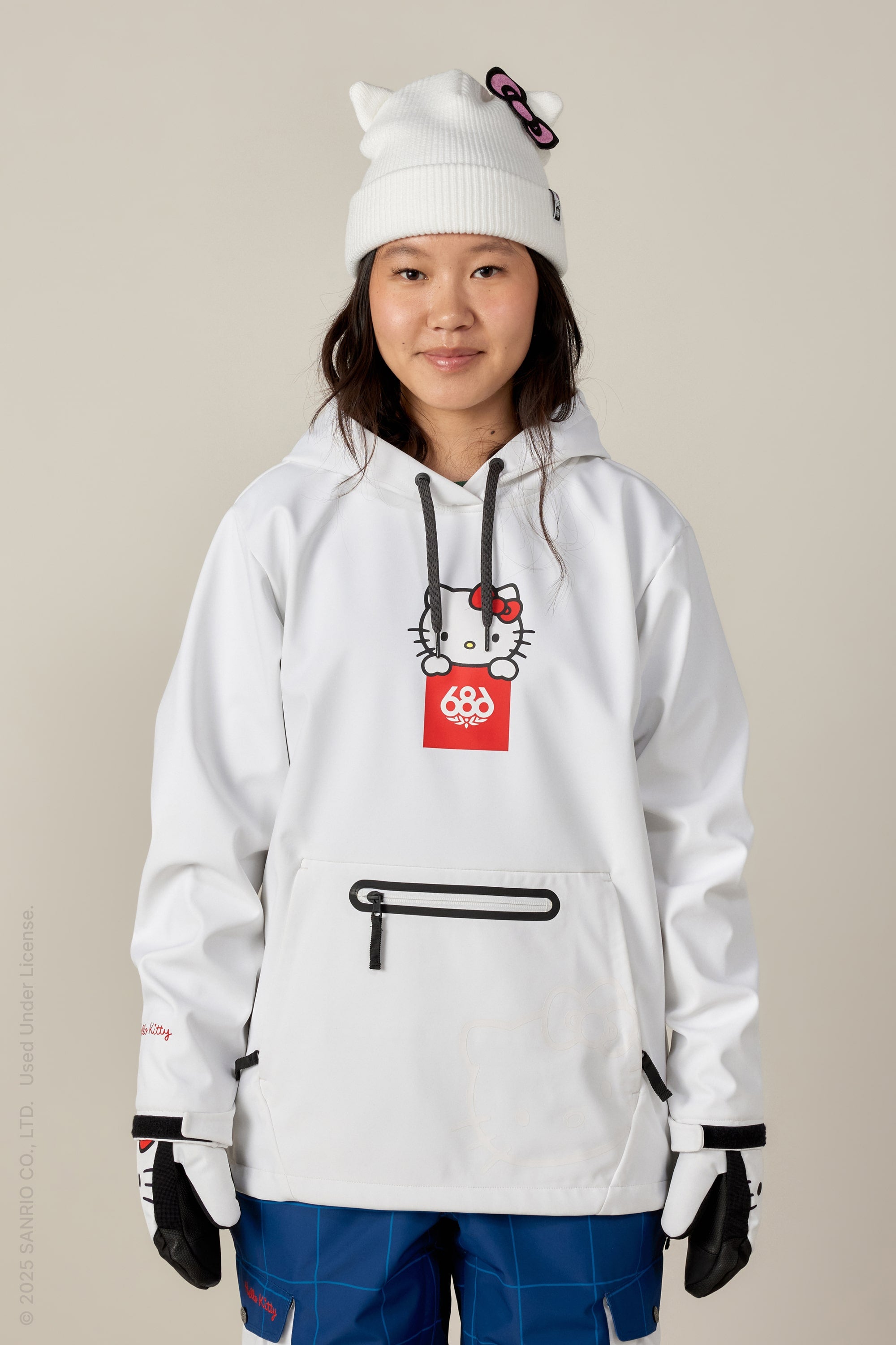 A woman faces the camera wearing a 686 Women's Waterproof Hoody in white with a Hello Kitty graphic, paired with a white Hello Kitty beanie, blue snow pants, and black-and-white gloves.