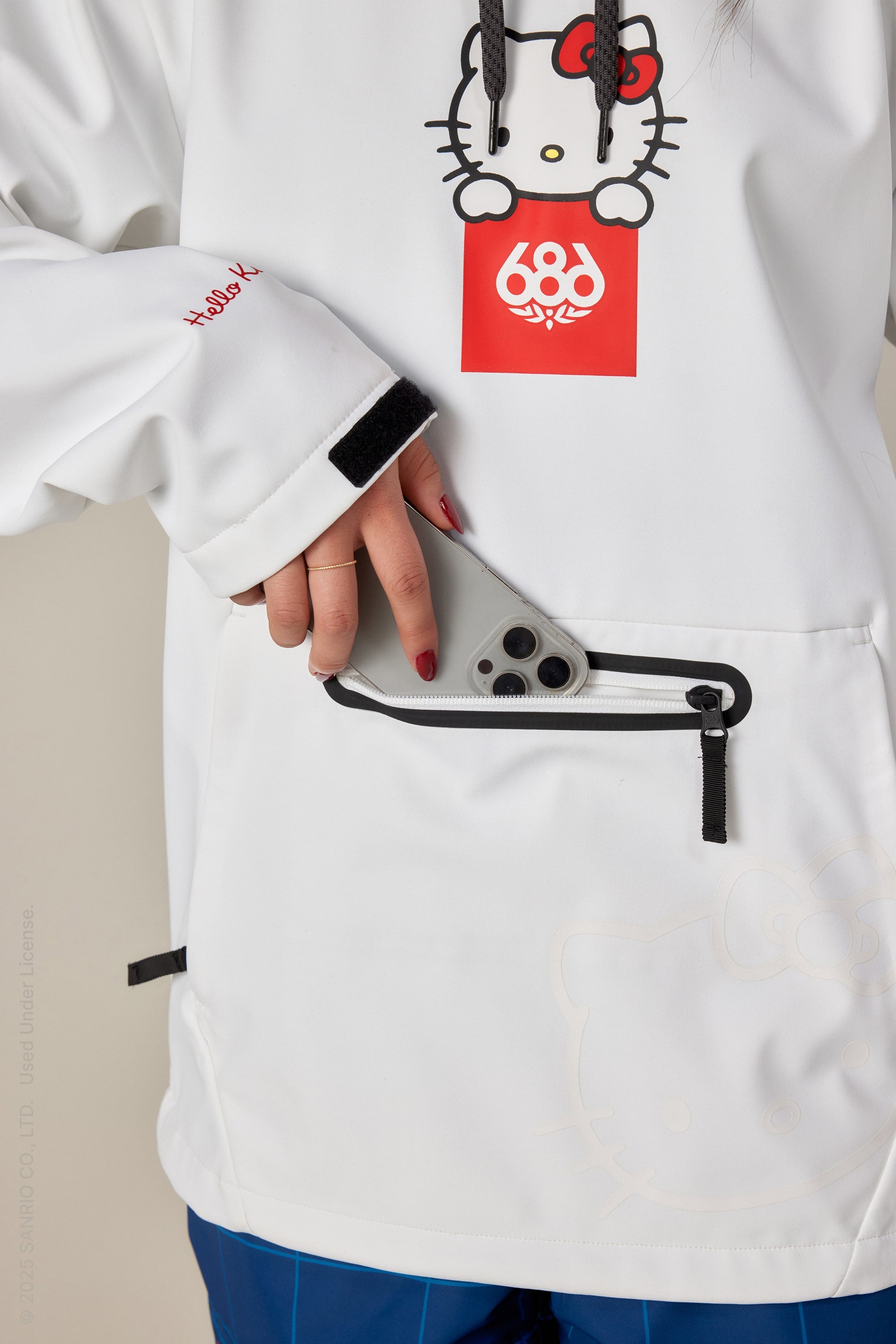 A woman in a 686 Women's Waterproof Hoody is slipping a gray smartphone into the front zippered pocket, with only the lower part of her face and torso visible.