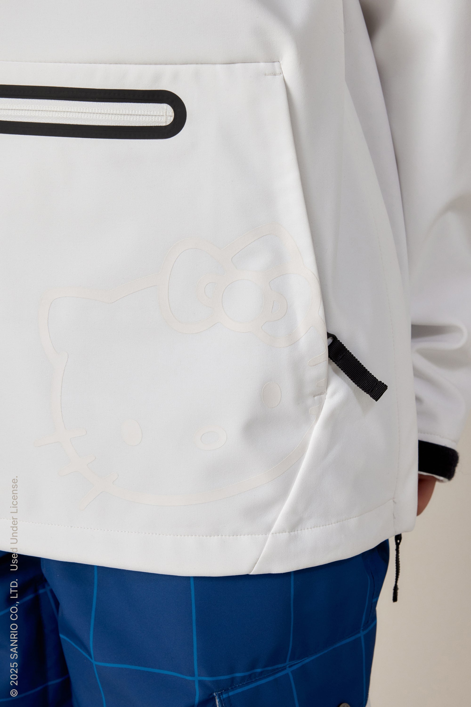 A person is wearing the 686 Women's Waterproof Hoody by 686, a white jacket with a subtle Hello Kitty face on the pocket, paired with blue grid-patterned pants. Their right arm is partially visible.