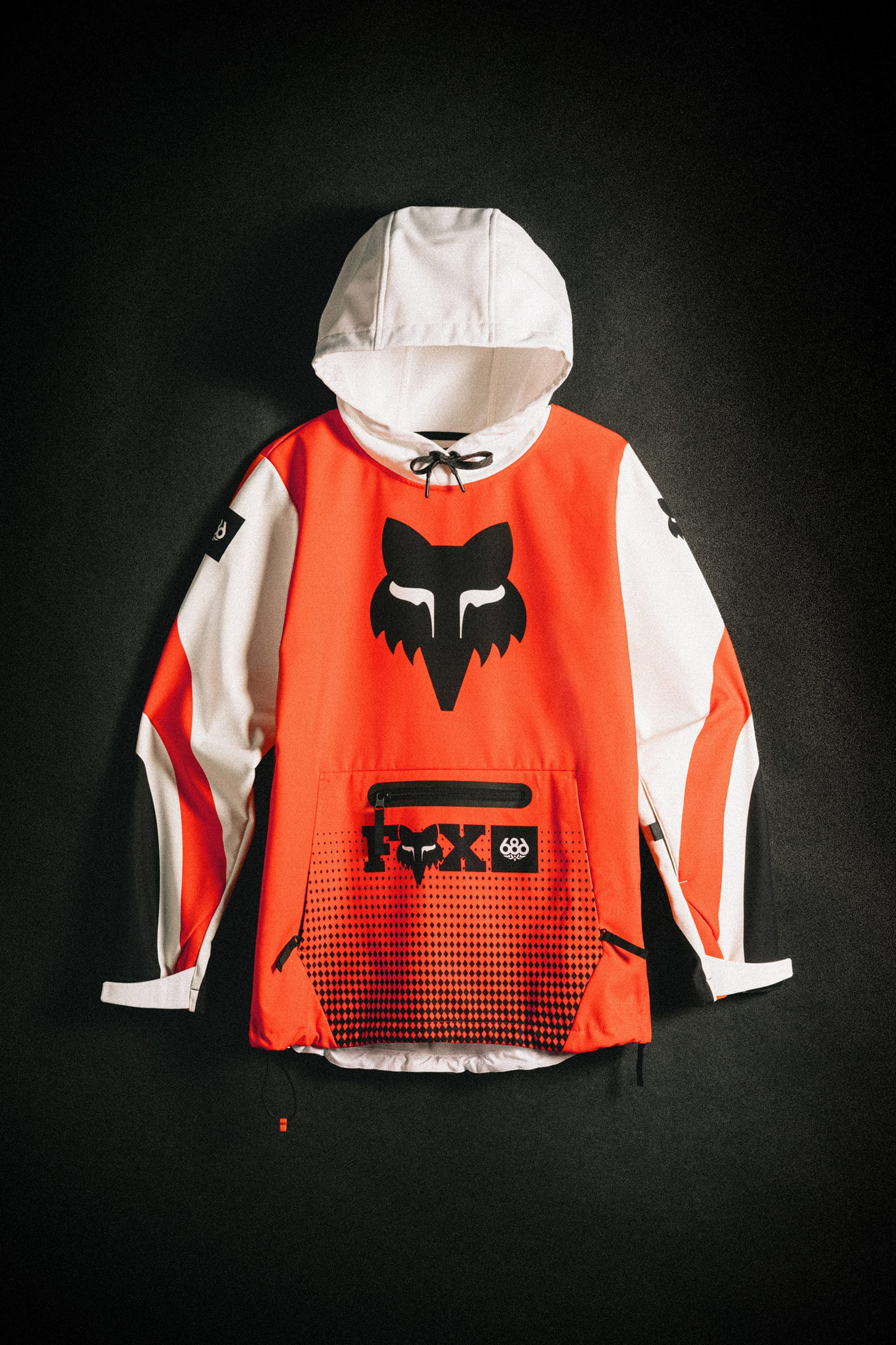 The 686 Women's Fox Racing 97 Waterproof Hoody features a white hood and sleeves, bright orange front and back, black Fox logo on the chest, black/red graphic pocket, and black drawstrings, shown laid flat against a dark background.