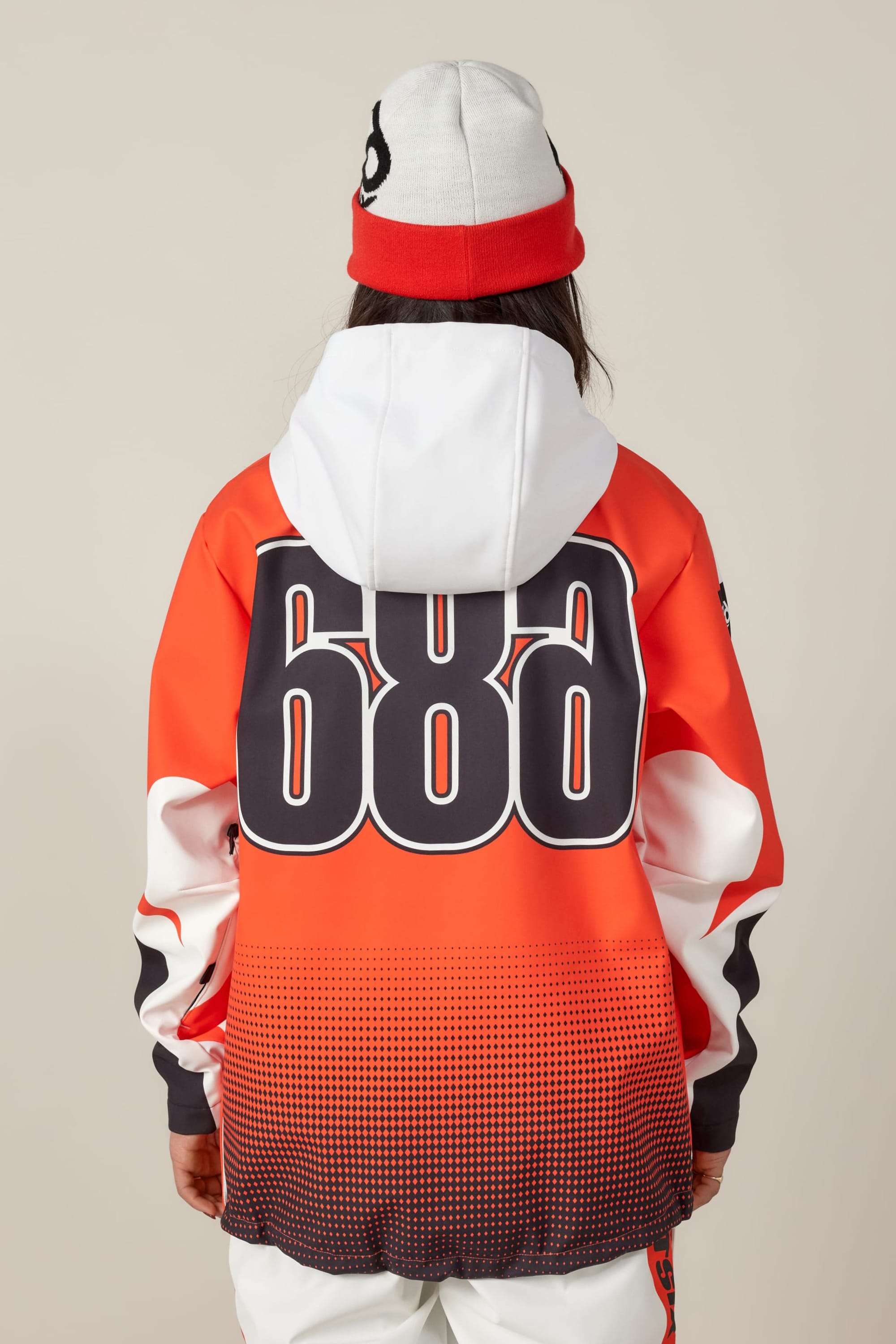 A person faces away, wearing the 686 Women's Fox Racing 97 Waterproof Hoody in orange with bold black "686" numbers on the back, paired with a white and red beanie. The background is plain and neutral.