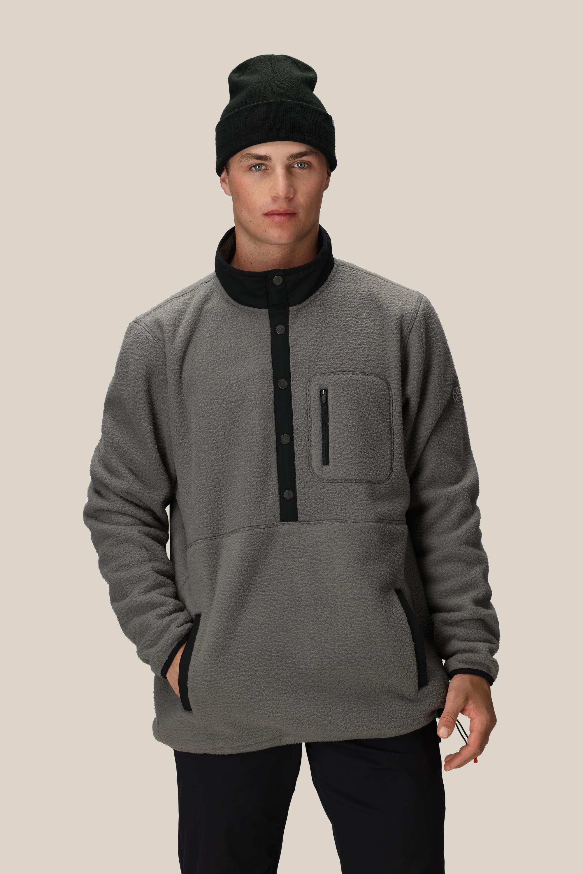A young man wearing a 686 Mens Tioga Fleece Pullover—featuring a button collar and chest pocket—stands with one hand in his pocket against a plain beige background.