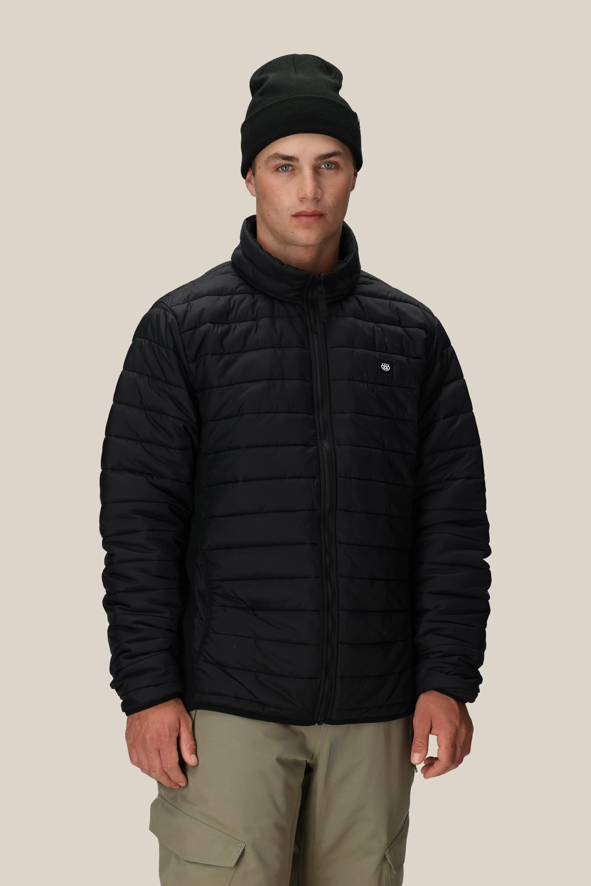 A person wearing the 686 Men’s Thermal Puff Jacket in black, paired with a black beanie and beige pants, stands facing forward against a plain beige background.