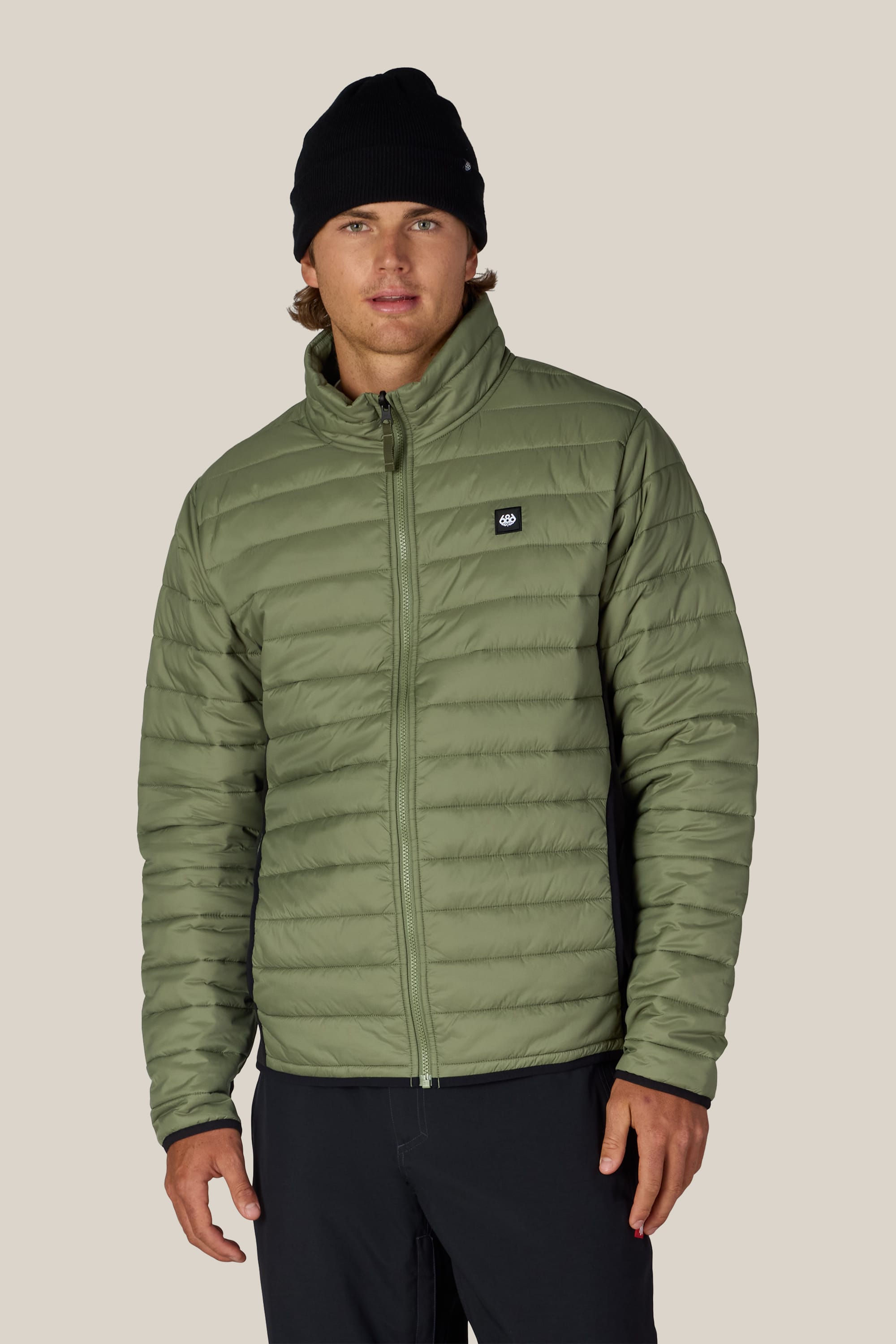 A man wearing a black beanie, black pants, and the 686 Men’s Thermal Puff Jacket by 686 in light green stands facing forward against a plain beige background.